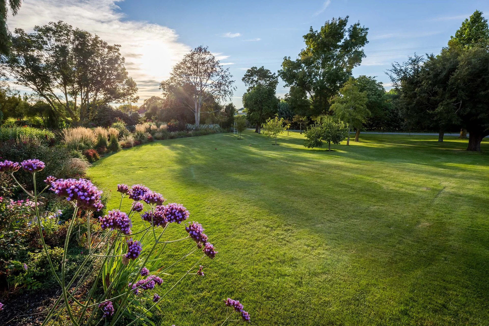 A lush green park with neatly mowed grass, colorful flowers in the foreground, and tall trees under a partly cloudy sky during sunset.
