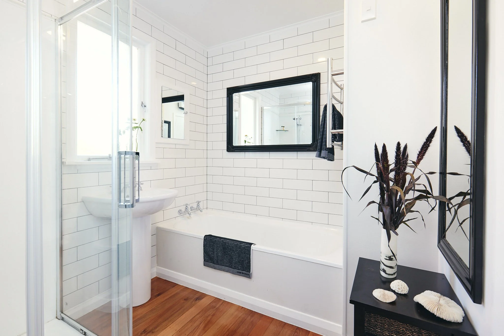 Modern bathroom with white tiled walls, a bathtub, a pedestal sink, a large black framed mirror, a small wall mirror, a window, and decorative plants and shells.