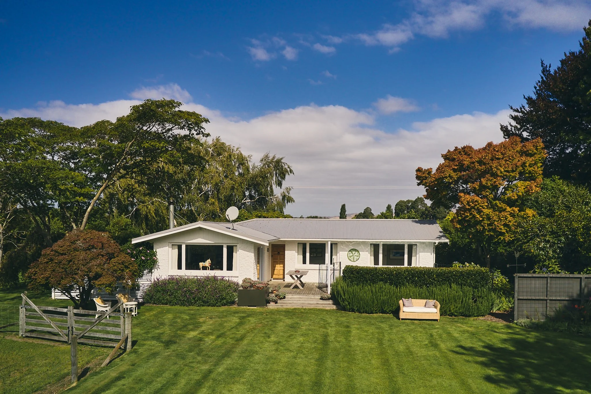 A white house with a gray roof and large front windows, surrounded by trees and a manicured lawn under a partly cloudy sky.