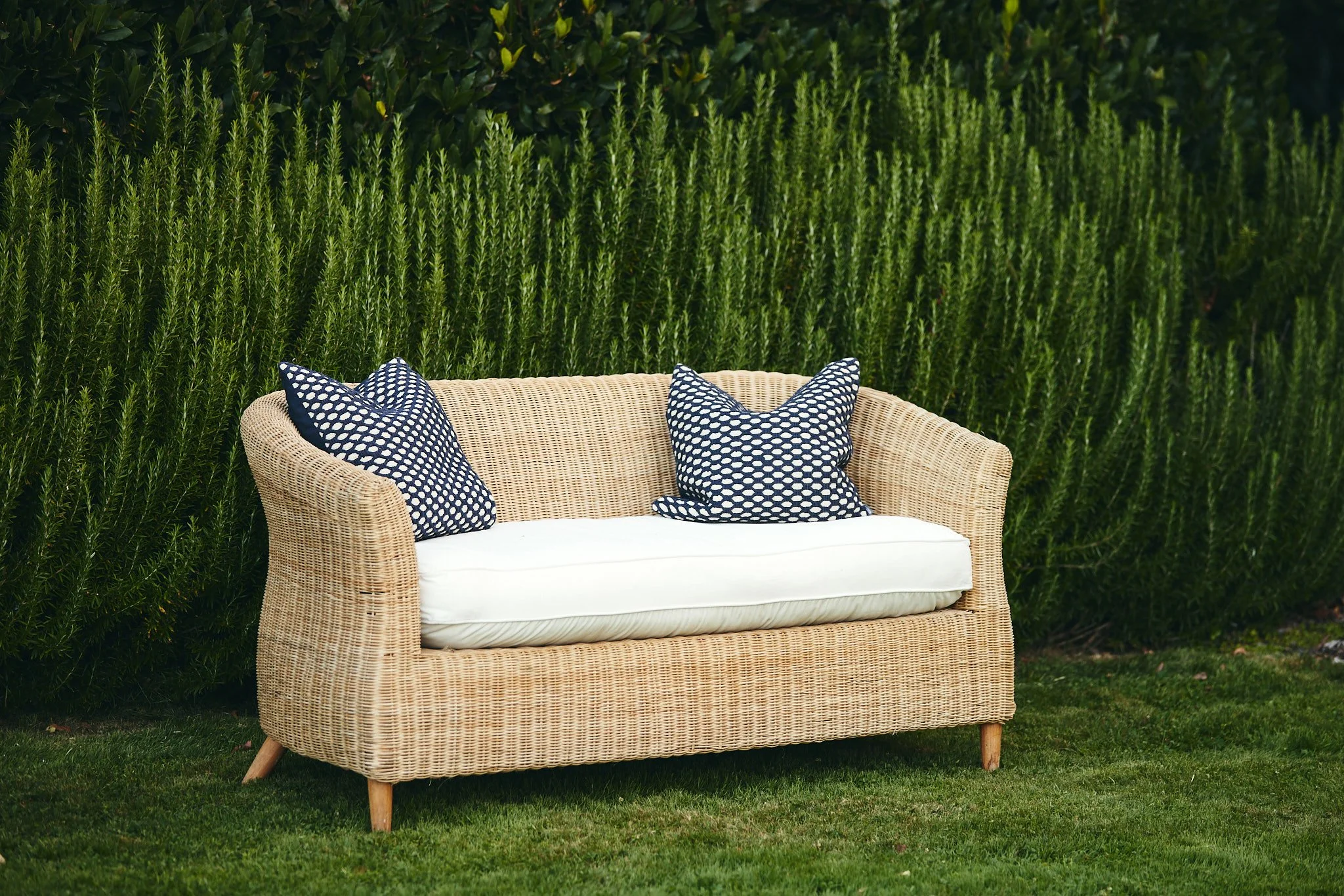 A wicker outdoor sofa with white cushion and two navy blue patterned pillows, placed on grass in front of dense green bushes.