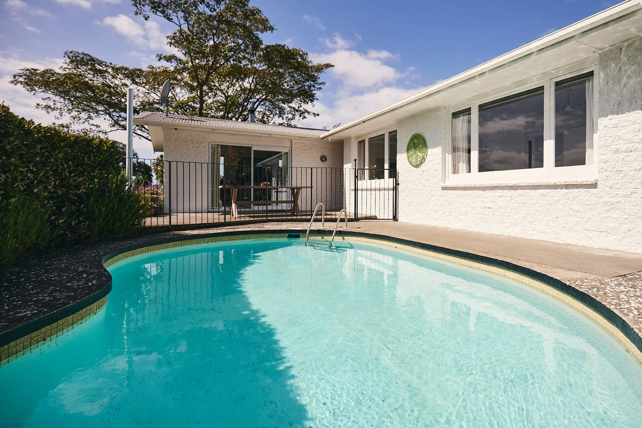 A backyard with a swimming pool, a white house with large windows, and a patio area with outdoor furniture under a partly cloudy sky.
