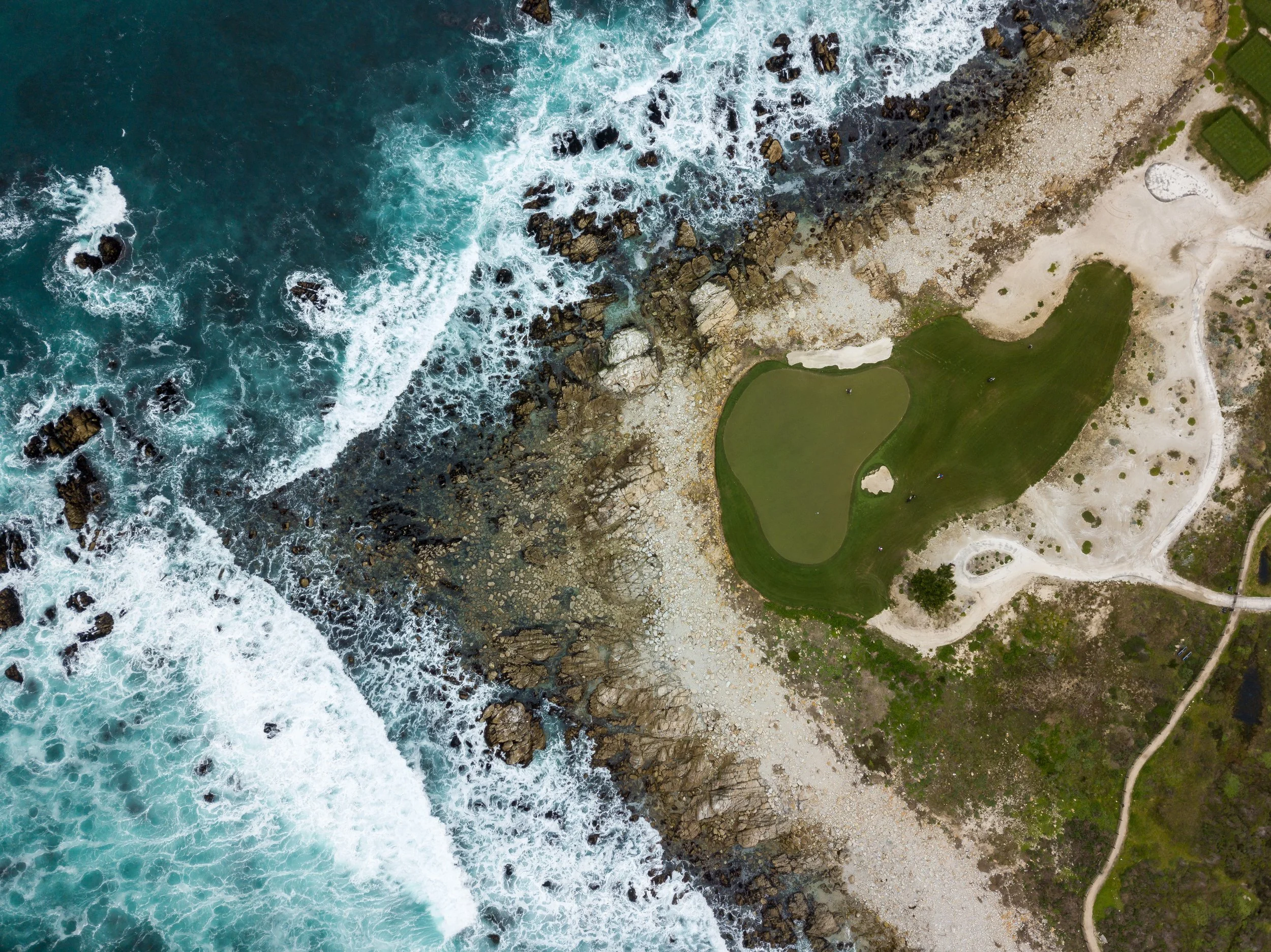 An aerial view of a rocky shoreline with waves crashing against the rocks, adjacent to a grassy area with sand and a golf course.