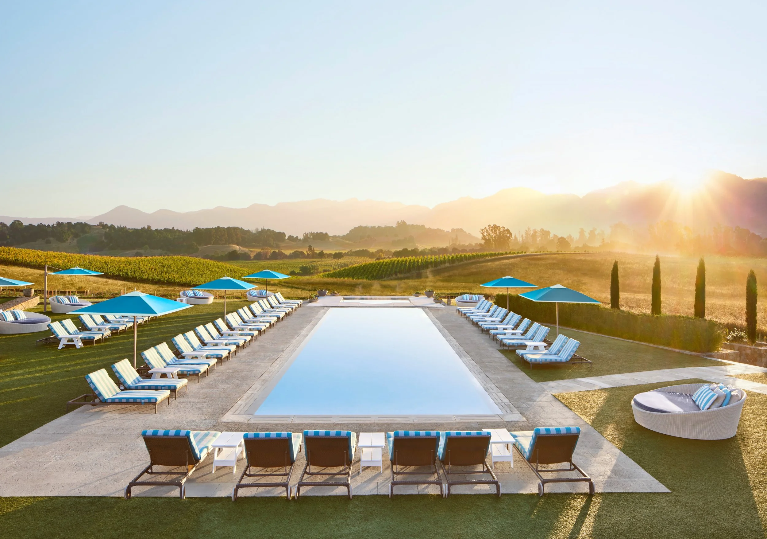 A luxurious outdoor pool area with lounge chairs and umbrellas, overlooking a scenic landscape and mountains at sunset.