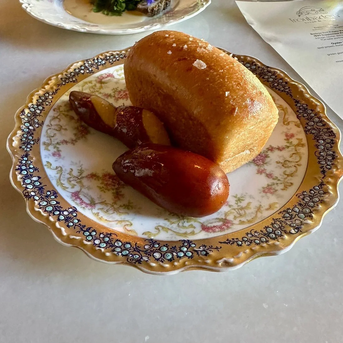 A plate with a large loaf of bread, two roasted sausages, and a potato wedge. The plate has a decorative floral border.