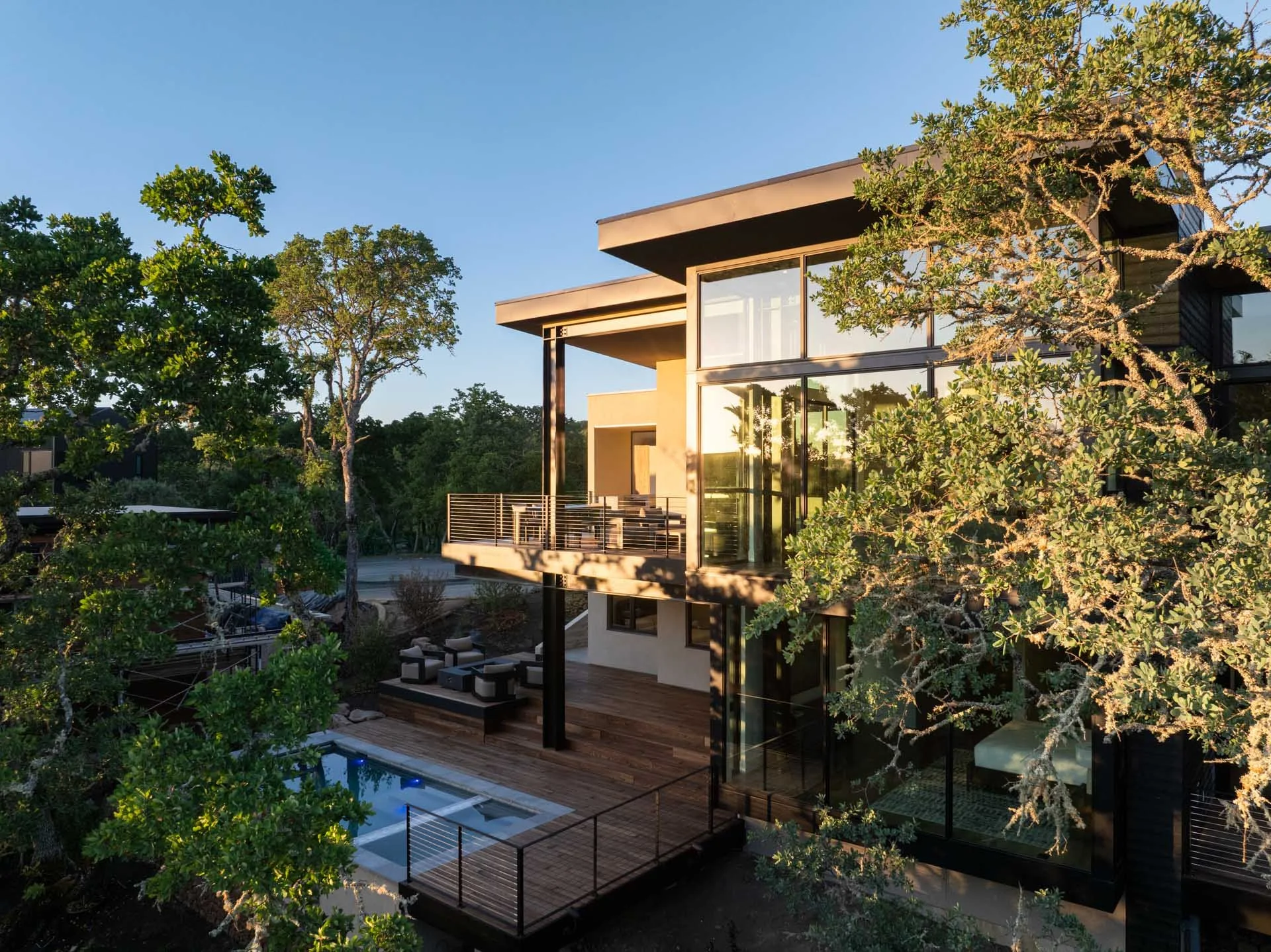 Modern multi-story house with large glass windows and balconies, surrounded by trees, during sunset.