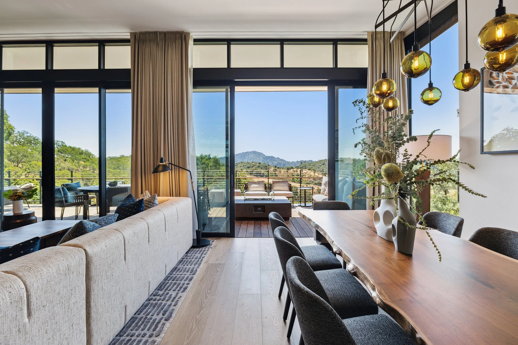 A modern living room with large floor-to-ceiling windows, beige curtains, a cream sectional sofa, a wooden dining table with gray chairs, a vase with greenery, and hanging yellow glass pendant lights, overlooking a balcony with outdoor seating and a hilly landscape in the background.