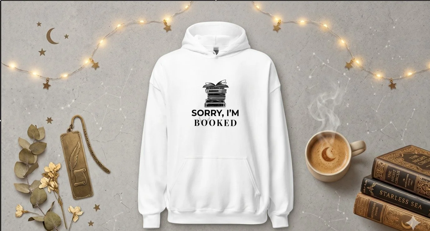 White hoodie with a stack of books graphic and the text 'Sorry, I'm Booked' on a flat surface surrounded by a cup of coffee, gold books, dried leaves, a book charm, and decorative string lights and stars on a grey background.