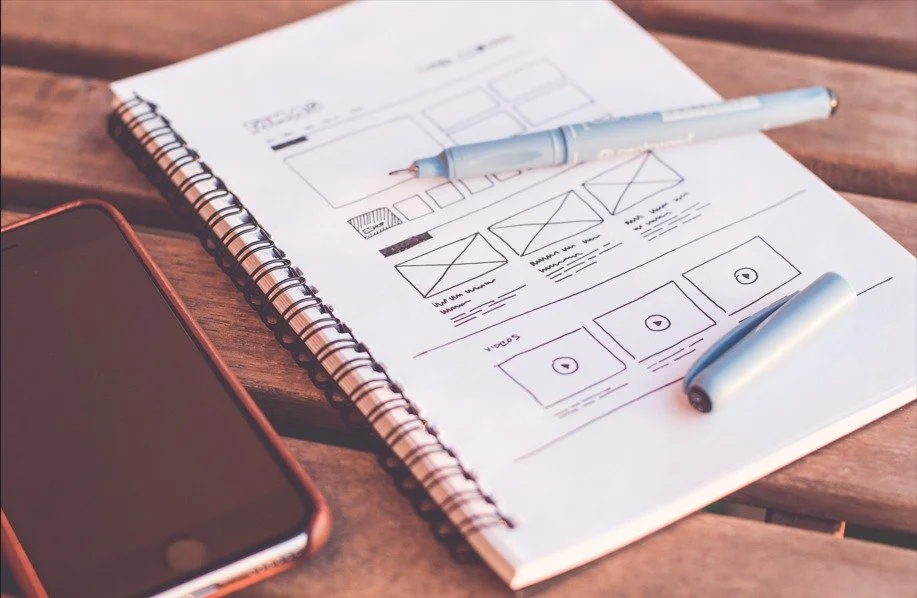 Sketch of a website or app layout on a spiral notebook with a pen, two video thumbnails, and a smartphone on a wooden surface.