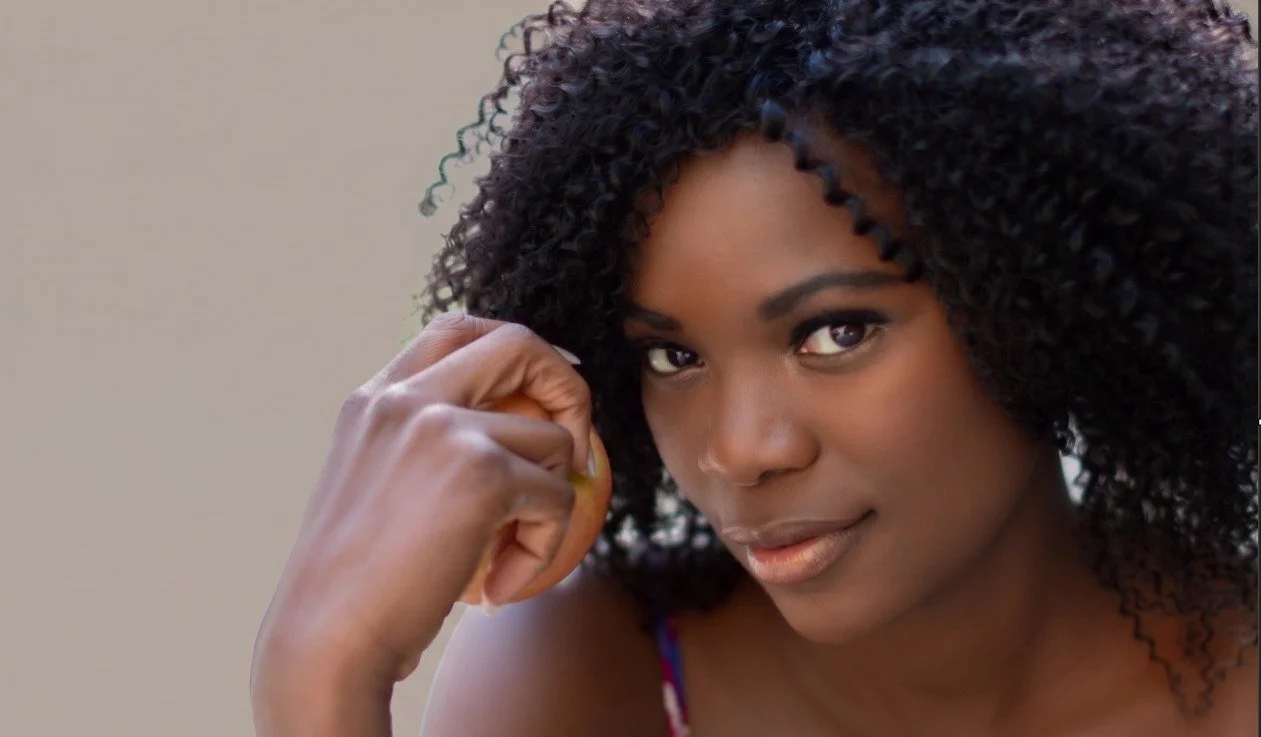 Close-up of a woman with dark skin and curly black hair, smiling softly, with her hand resting near her face.