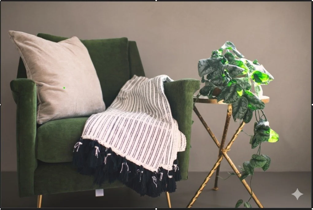 A green upholstered armchair with a beige pillow and a black and white striped blanket, placed next to a small wooden stand with a leafy green plant.