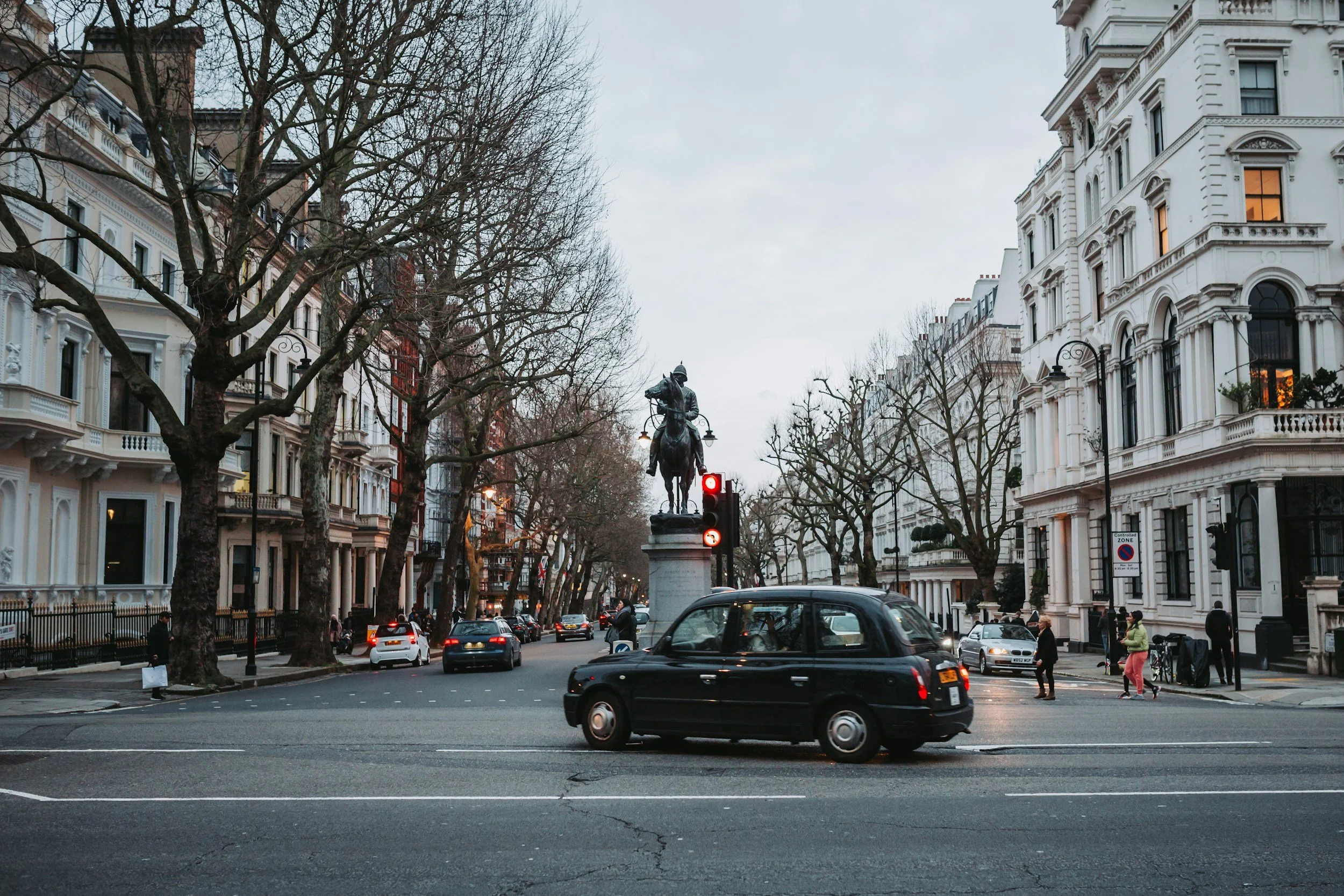 City street scene with a black taxi driving past a traffic light, a statue of a man on horseback in the center, and white historic buildings with leafless trees lining the street.