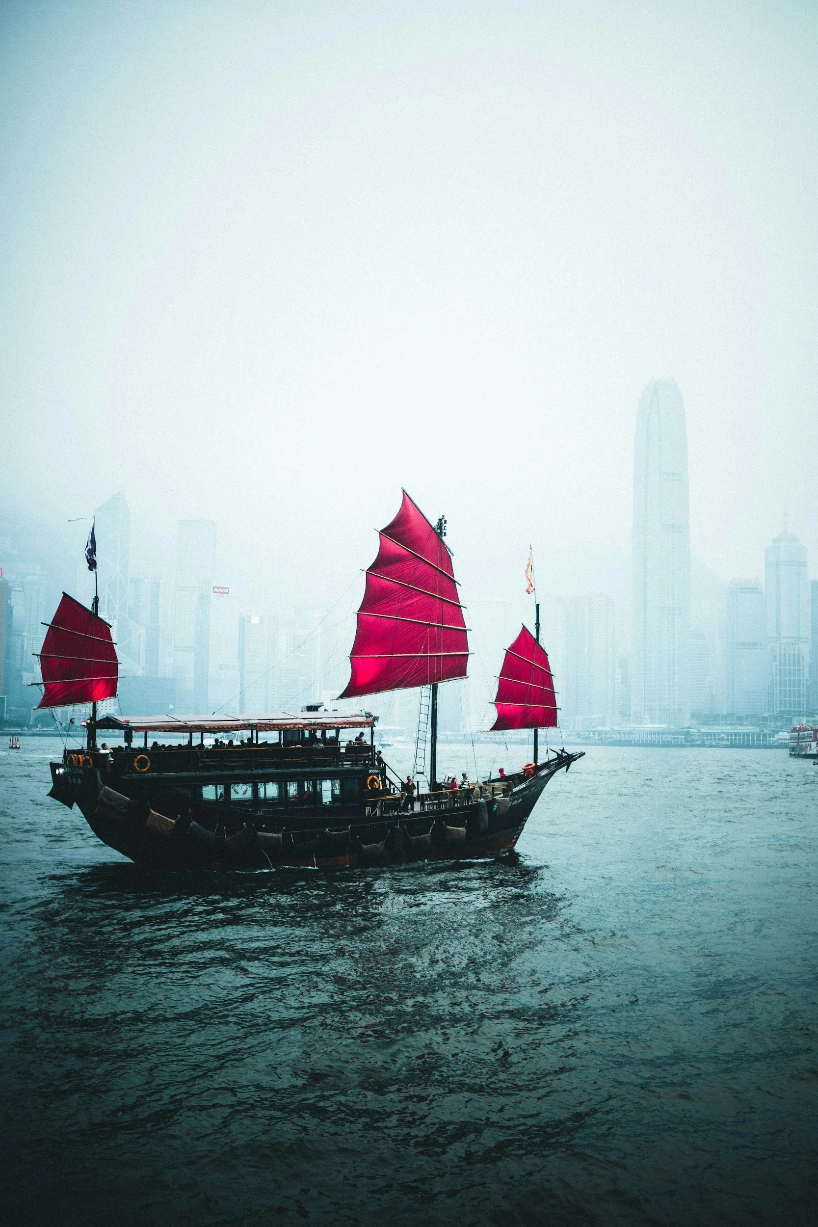 A traditional Chinese junk boat with red sails navigates across Victoria Harbour in Hong Kong, with the city skyline shrouded in fog in the background.