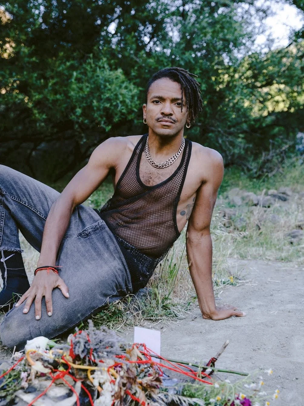 a black nonbinary person with short locs and a medium silver chain necklace - leans on one arm in a loose mesh tank, staring deeply at the camera behind a blurred mix of various offerings at the center of a rocky altar. 