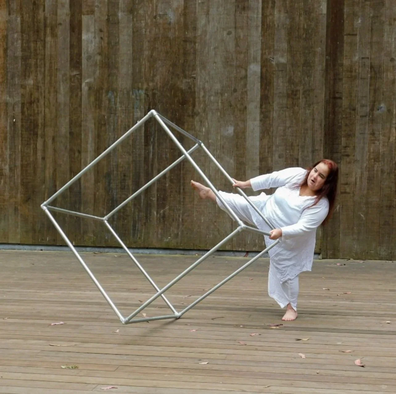 a full body shot of Maia in all white balancing on one leg, as she interacts with a large cube sculpture made of pipes