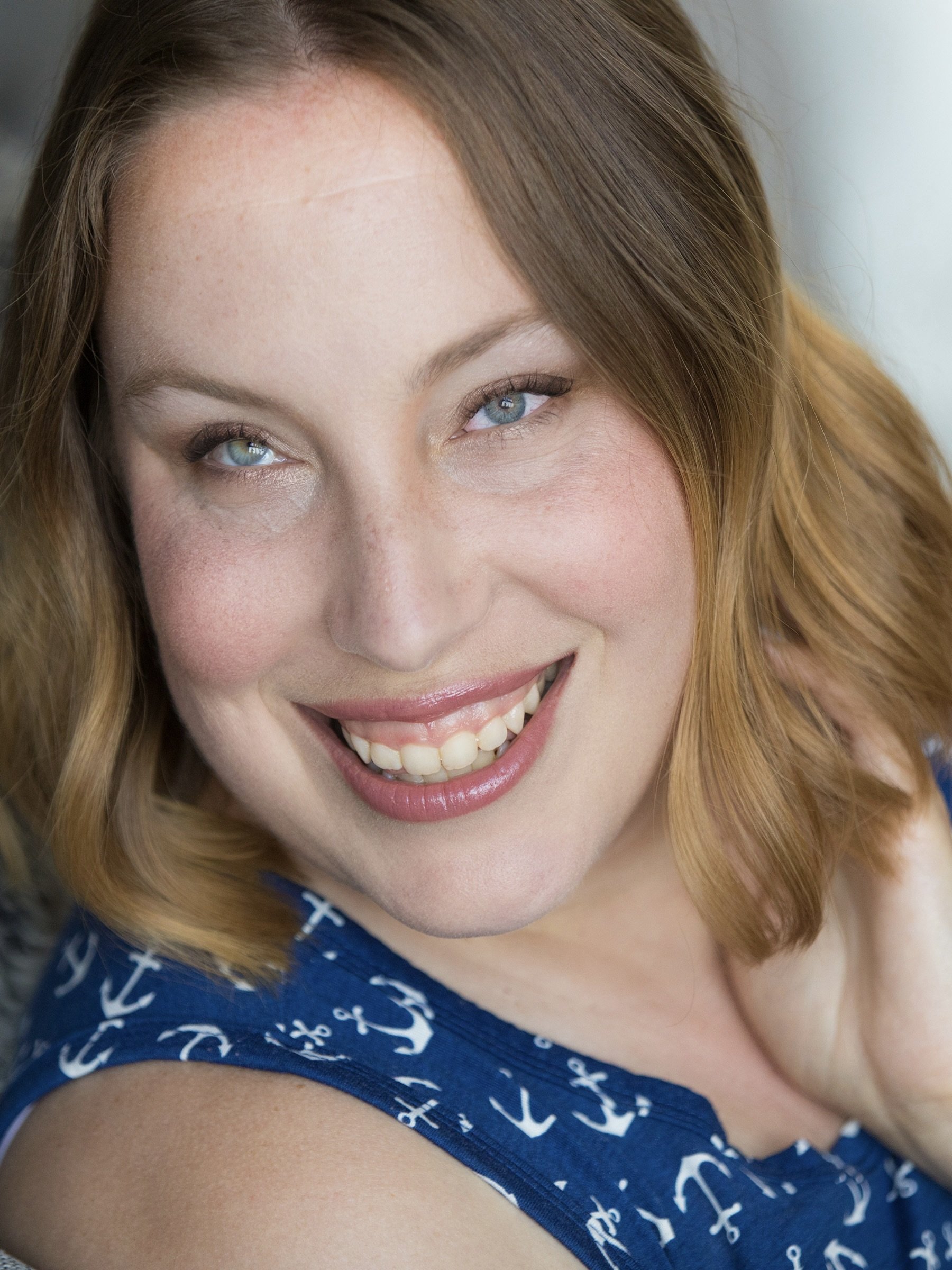 A headshot of Tiffany Taylor, she/her, a blind white woman in her 30s. She has chin length blond wavy hair, and green eyes. She is wearing a blue shirt with anchor designs and is smiling at the camera.