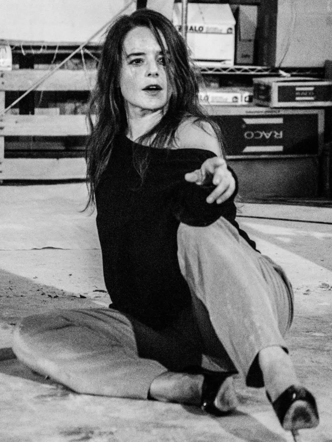 A black & white photo of a white woman with long dark hair, grey slacks, and an oversized off-the-shoulder shirt. She is sitting on the floor, hand on knee looking over her shoulder..