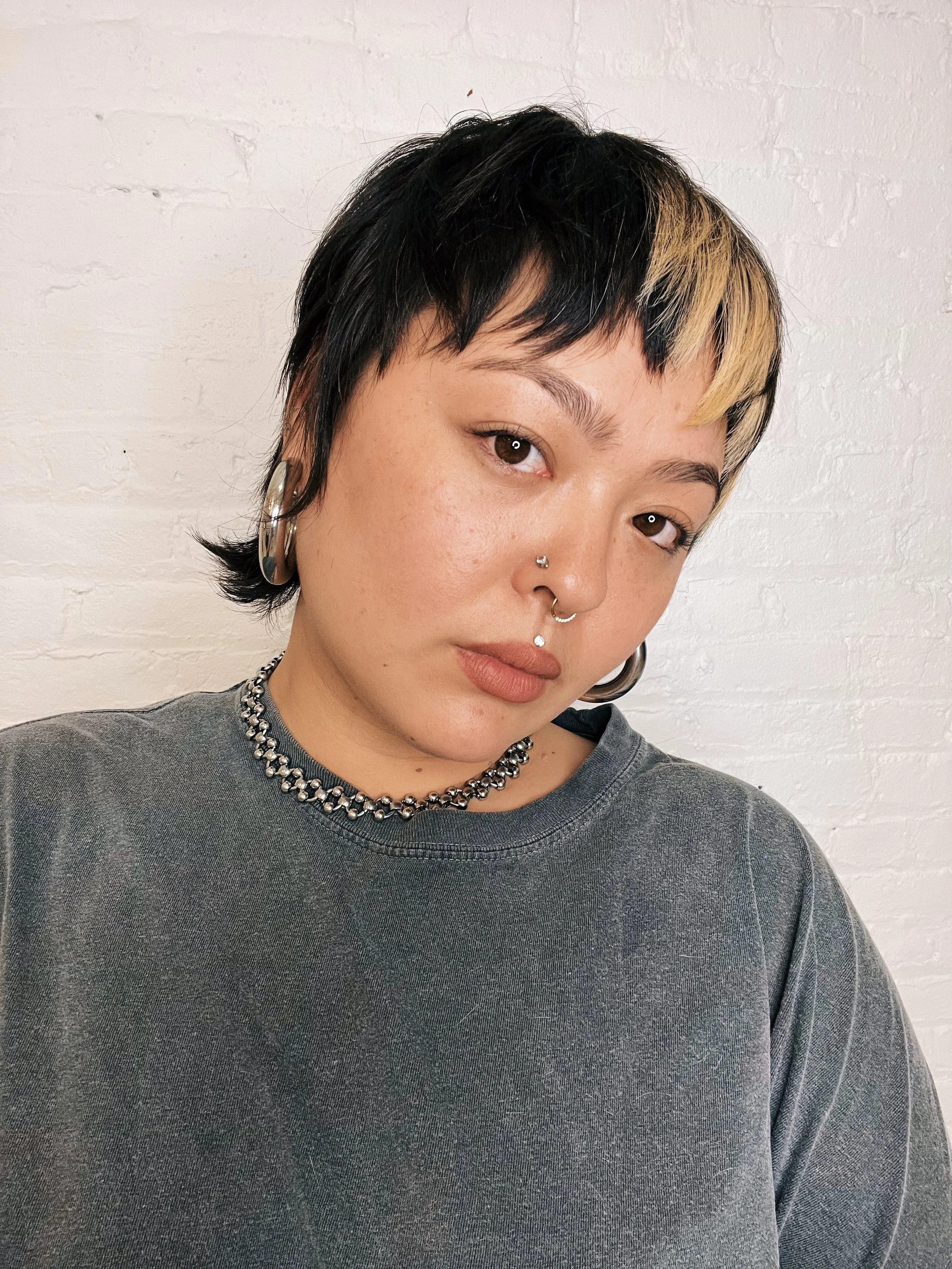 A headshot of Aiano from the chest up. They have short brown hair with blond bangs. They are wearing a grey shirt and a silver earrings and necklace.