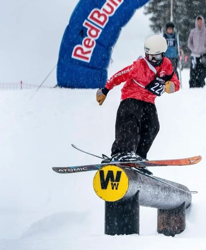 A young skier wearing a white helmet, red jacket, black pants, and goggles is performing a ski trick, jumping over a yellow sign with a black logo, on a snowy slope during a competition or event, with a Red Bull banner in the background.