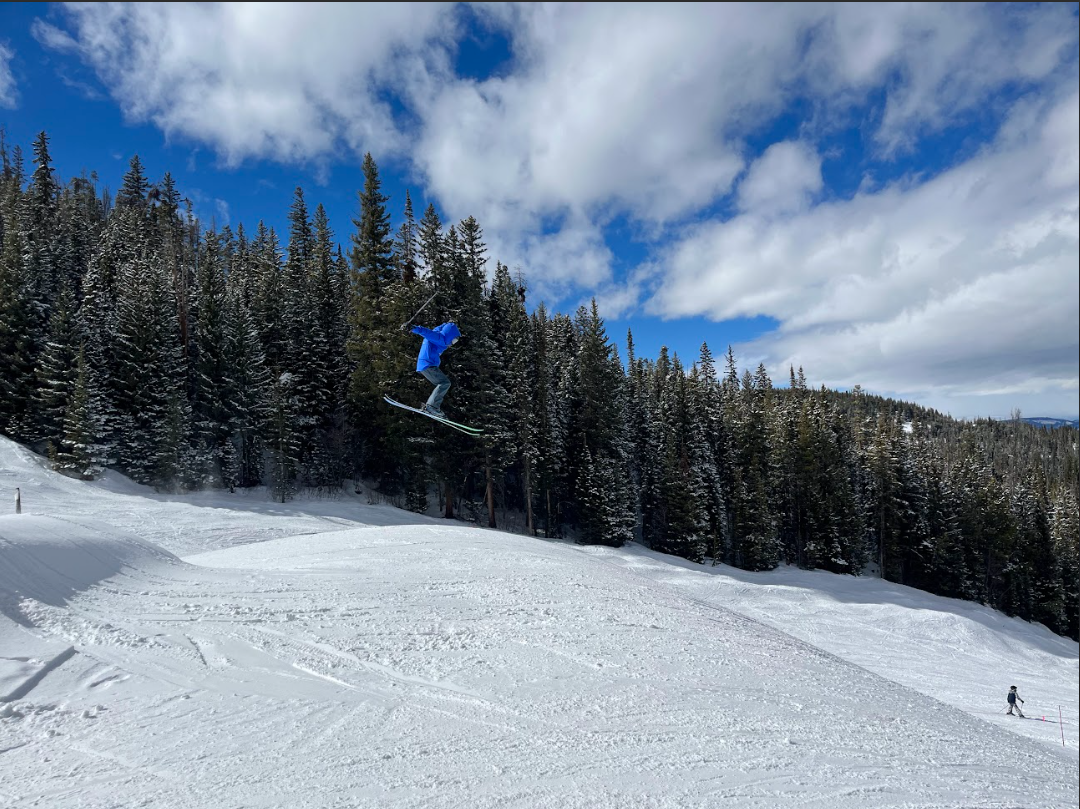 A person in a blue jacket is jumping in the air while skiing on a snowy slope, with a forest of pine trees and a partly cloudy sky in the background.