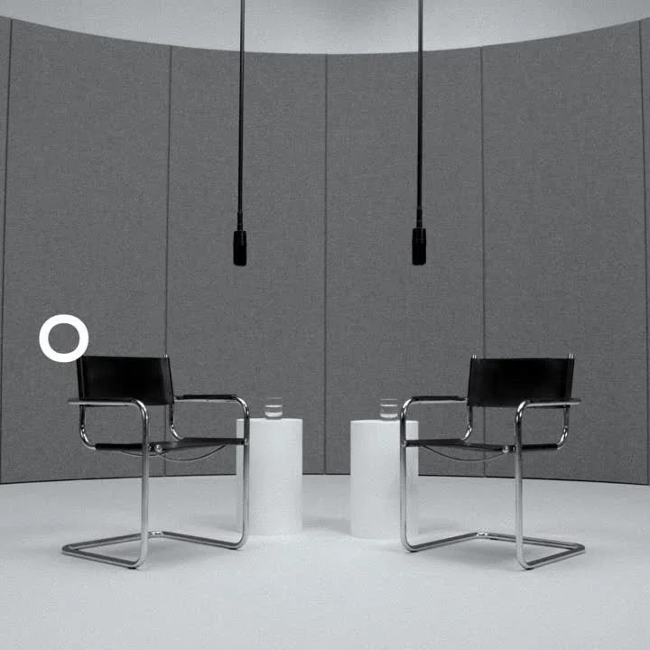 Two black chairs with chrome legs and armrests face each other in a minimalist, gray room with a curved wall, two black pendant lights hanging from the ceiling, and two cylindrical white side tables with glasses of water on top.