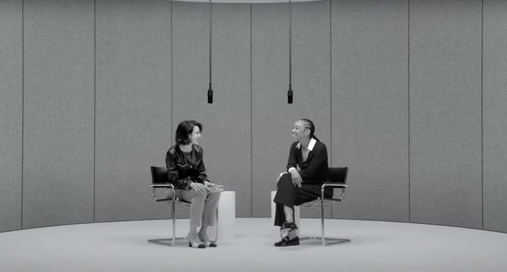 Two women sitting on chairs facing each other in a minimalistic room, engaged in conversation.