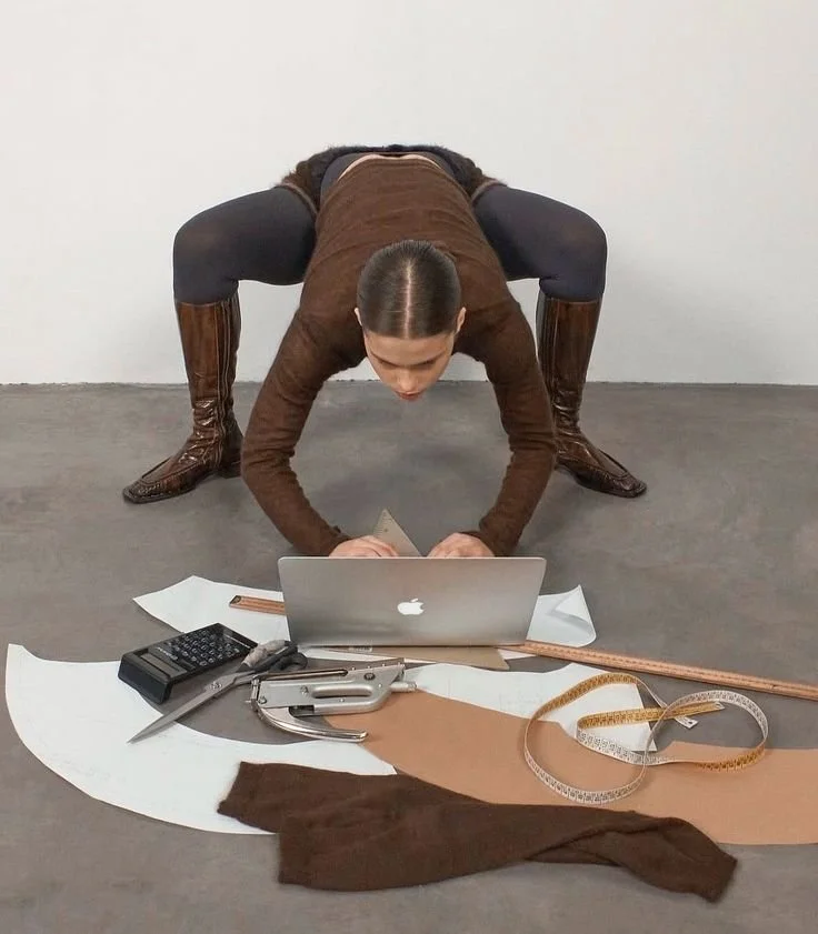 Person in a brown sweater and black leggings crouches over a laptop on the floor, surrounded by sewing patterns, fabric, and measuring tools.