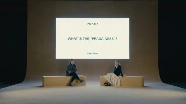 Two people sitting on stage in front of a large screen displaying the text, 'What is the "Prada-ness"?' with the Prada logo, during a presentation or talk.