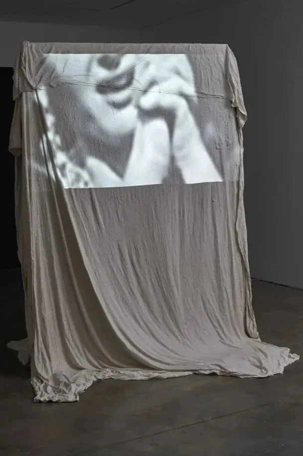 A black-and-white projection of a person with glasses, possibly an older man, is displayed onto a draped fabric surface.