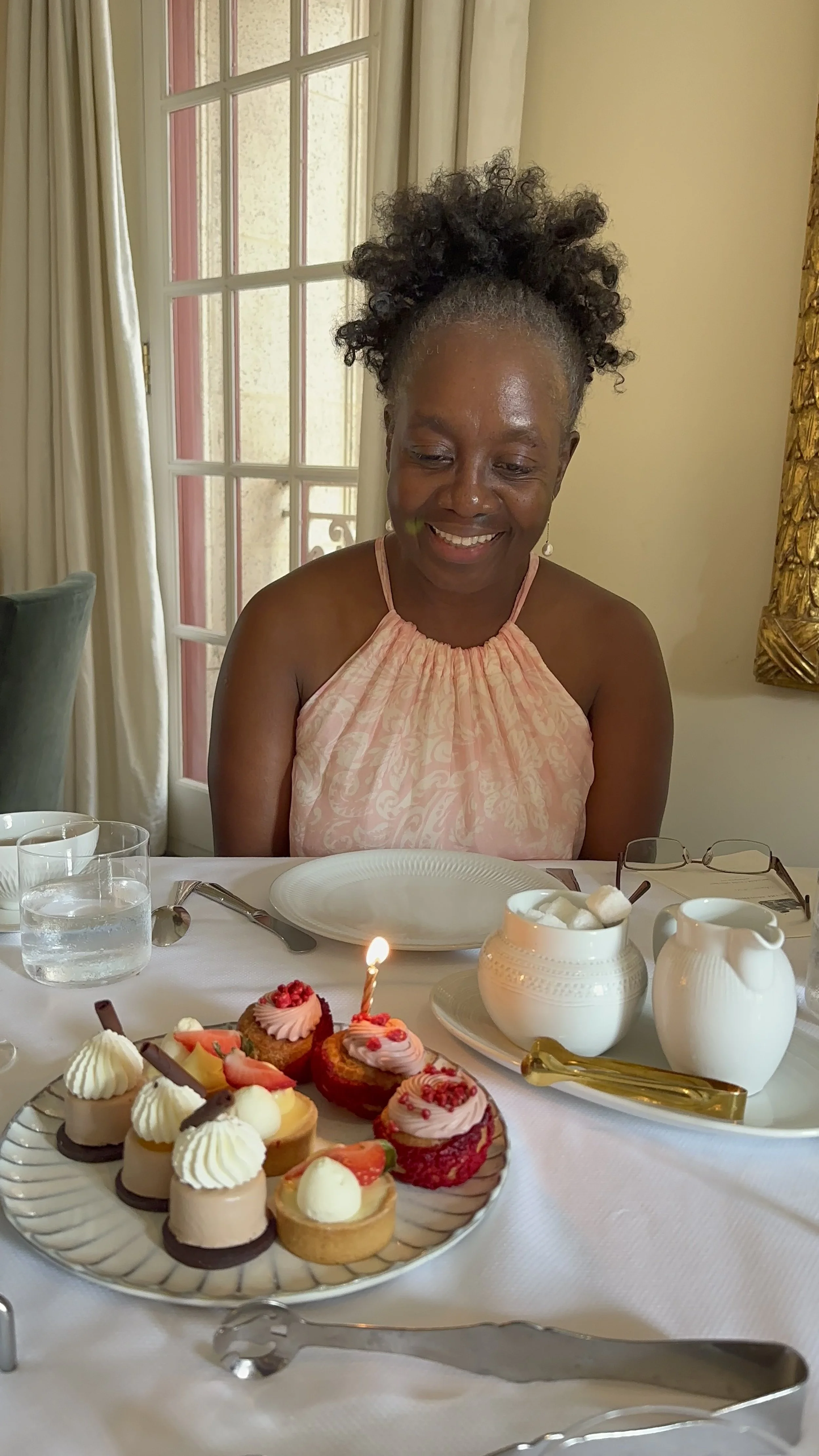A woman smiling at a birthday dessert table with various mini cakes and cheesecake bites, decorated with fruit and cream, with a lit candle on one of the desserts.