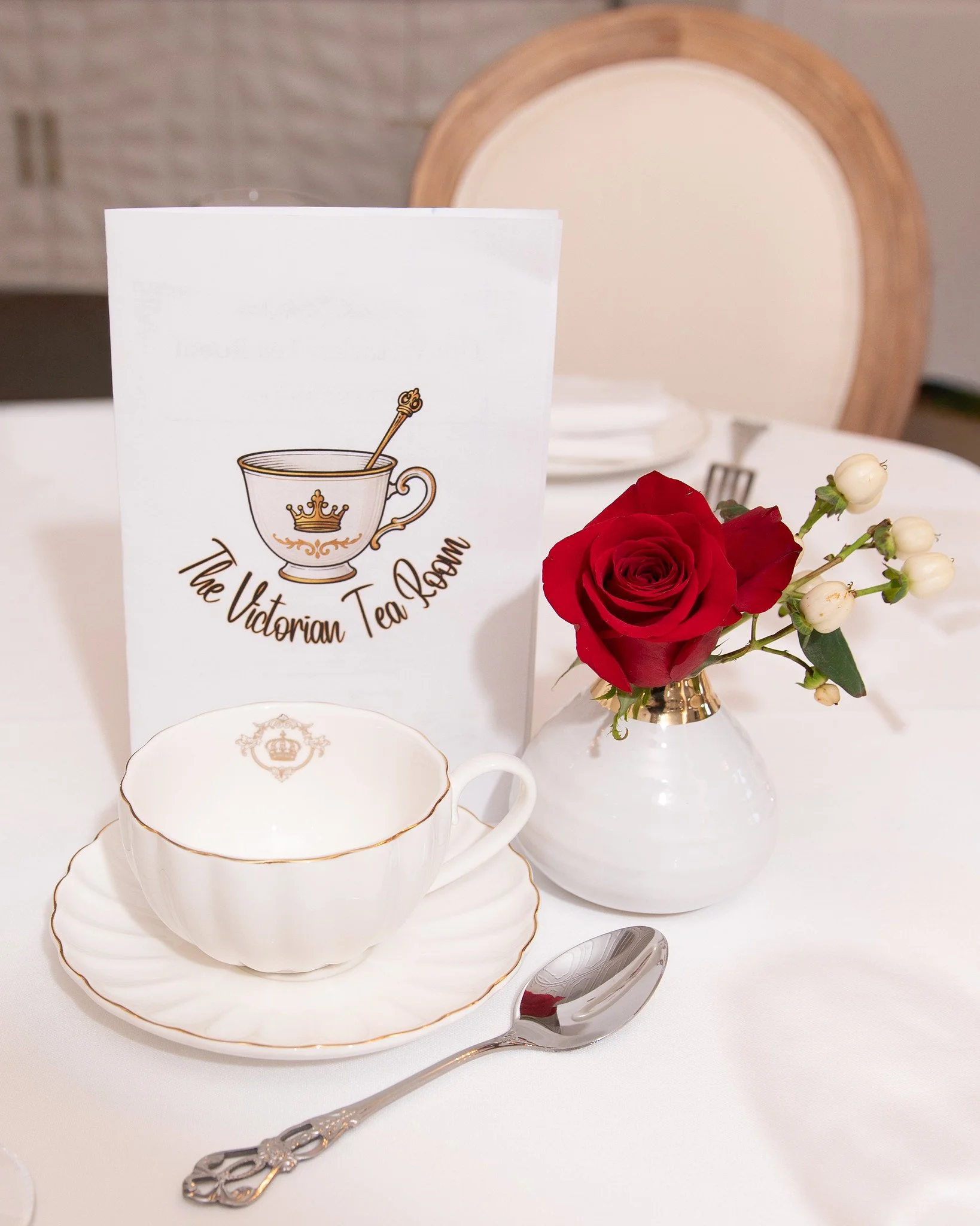 A table setting with a white teacup and saucer with gold trim, a silver spoon, a small vase with a red rose and white berries, and a menu card for The Victorian Tea Room, on a white tablecloth and a wooden chair in the background.