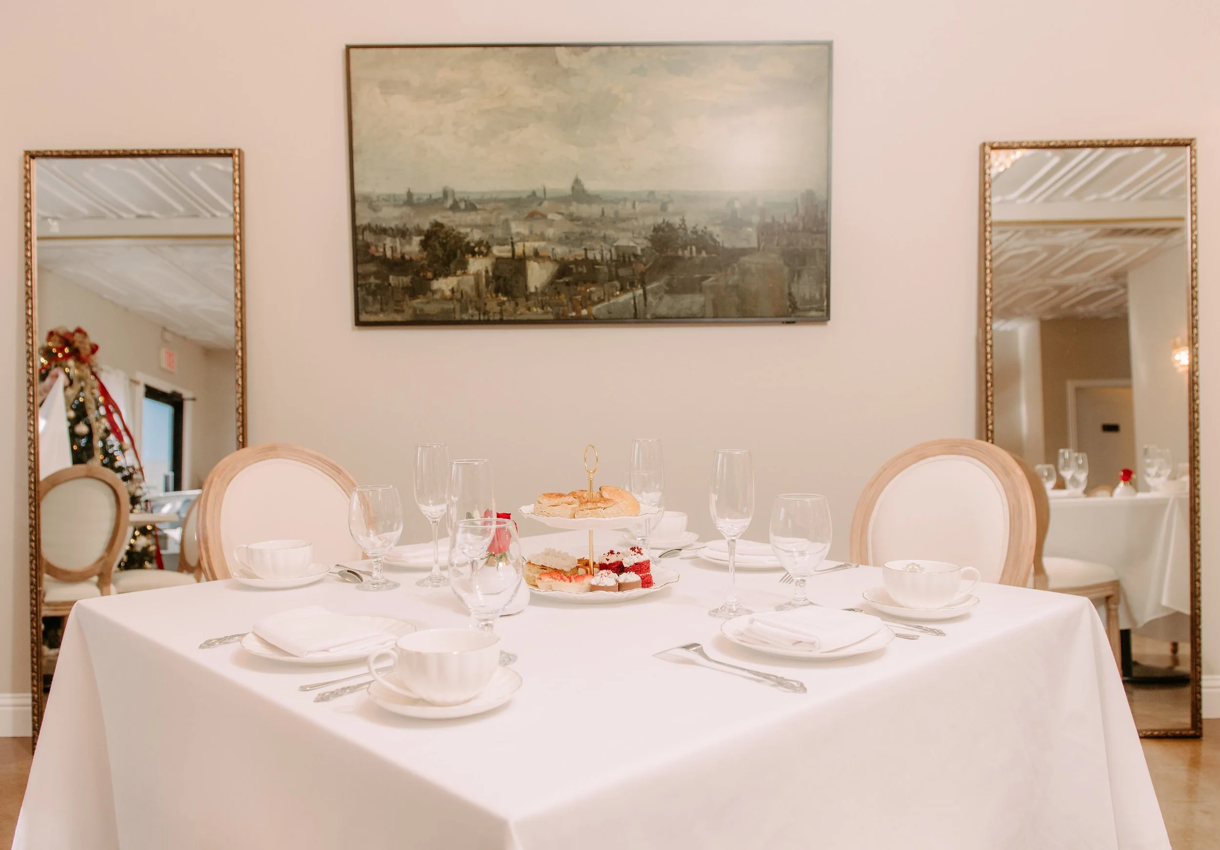 A dining table set for a meal, with white tablecloth, dishes, glasses, cups, silverware, a tiered serving platter with desserts, and chairs around it, in a decorated restaurant or event space with mirrors and artwork on the walls.