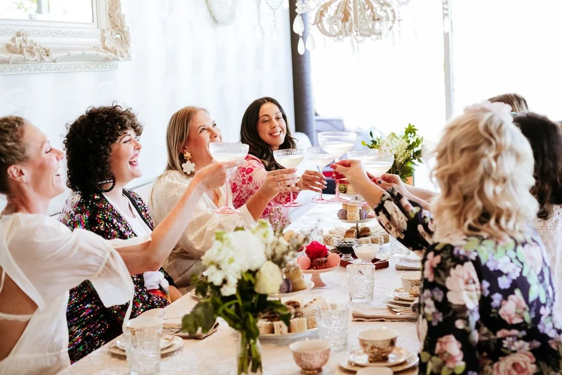 Group of women celebrating at a table with desserts and drinks, toasting with cocktail glasses, smiling and laughing.