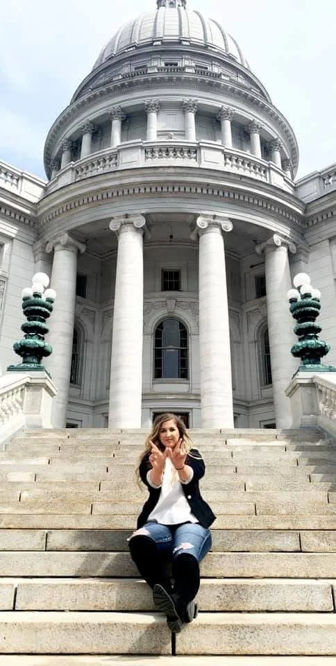 Victoria LaBrosse sitting on the steps of the Wisconsin Capitol and holding her hands in the shape of a W