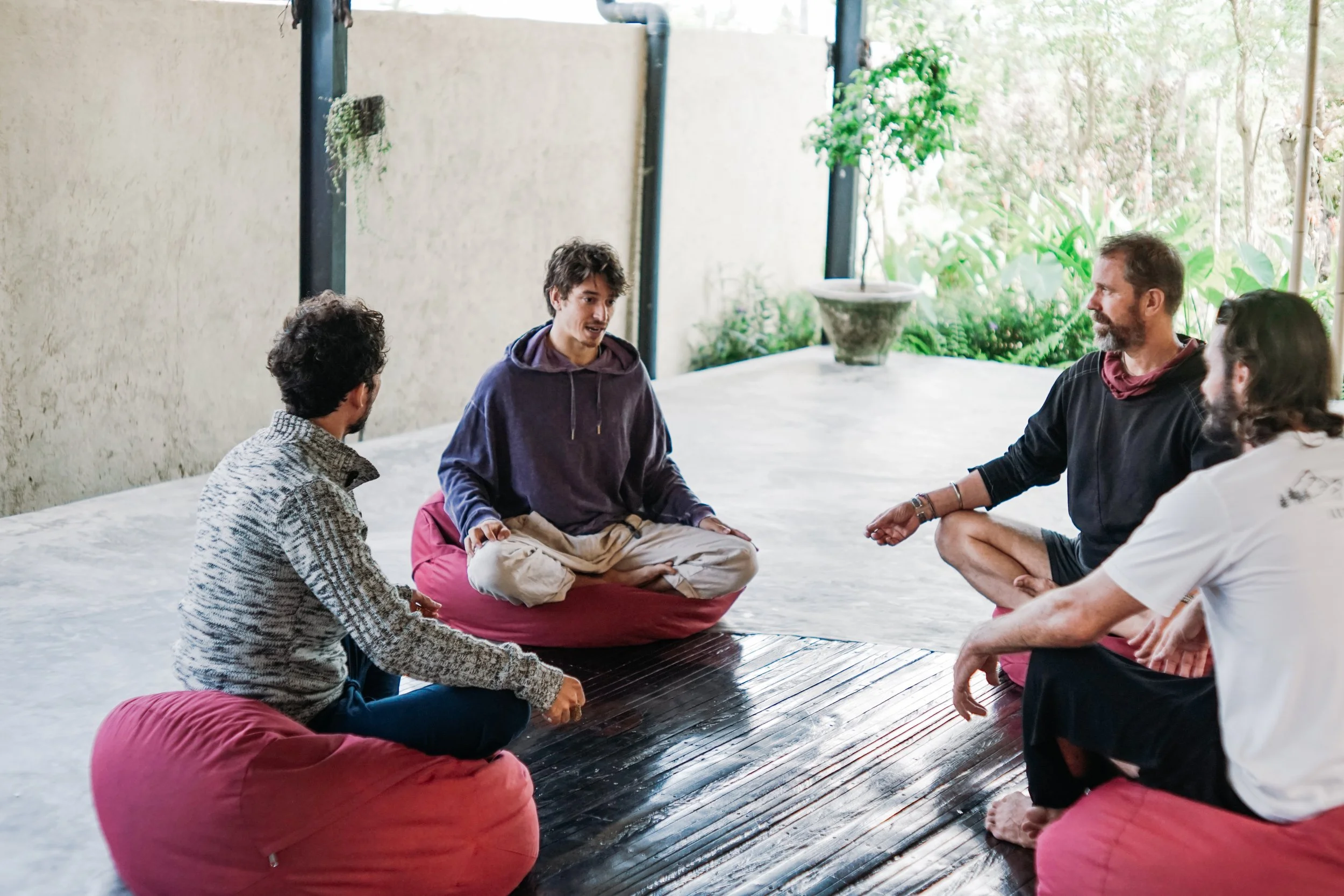 Five people sitting on cushions in a circle during a discussion, with greenery outside visible in the background.