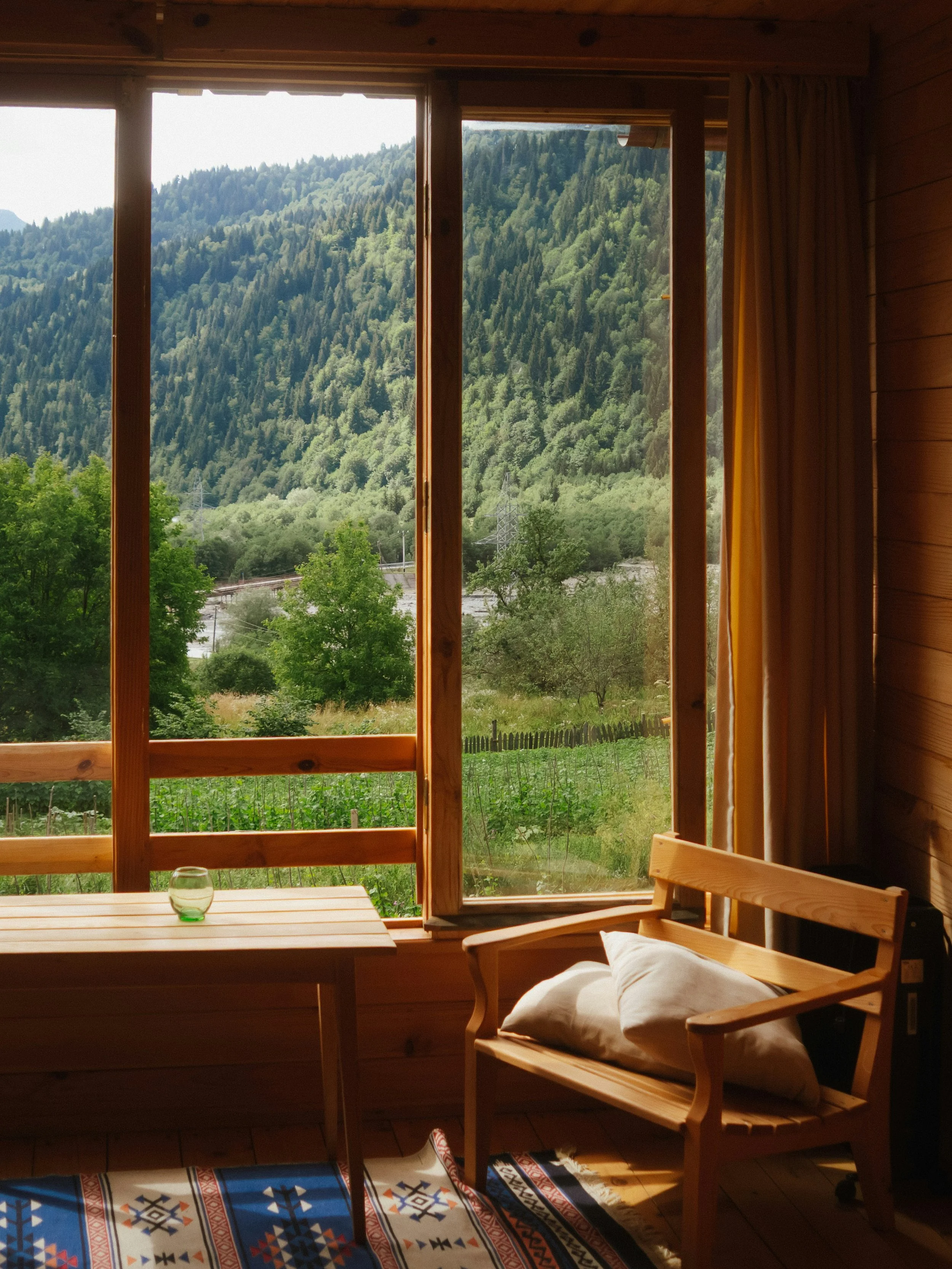 View of a lush green mountain landscape seen through a large window in a cozy wooden room, with a small wooden table, a chair with a cushion, and a colorful patterned rug on the floor.
