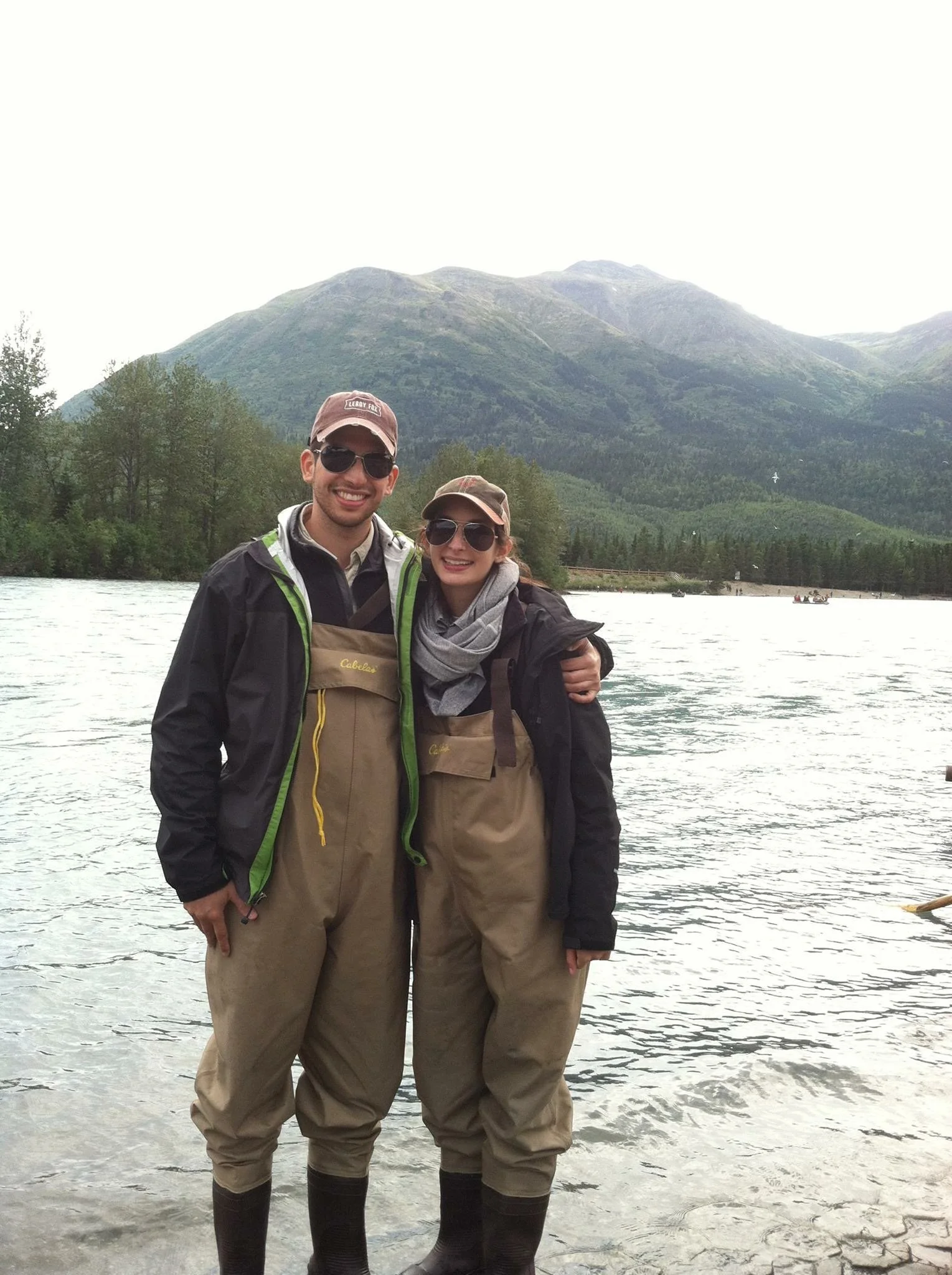 Two smiling people in outdoor fishing gear standing in shallow water near a mountain with trees and boaters in the background.