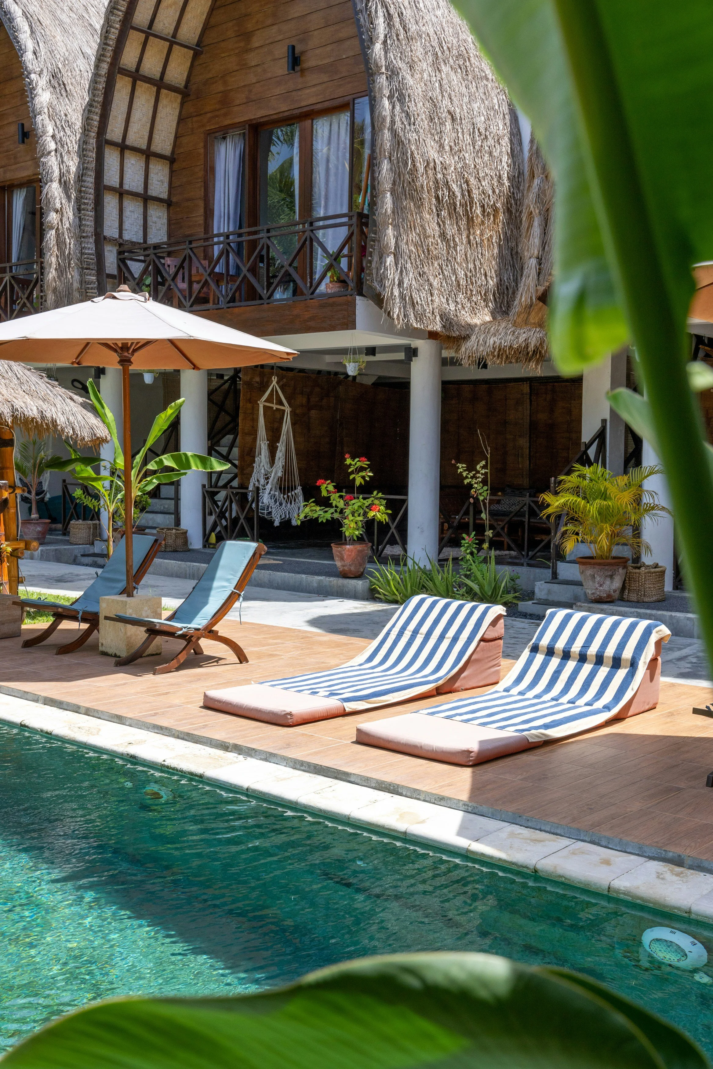 Poolside scene with lounge chairs, a large umbrella, tropical plants, a wooden building with a thatched roof, and a hammock, suggesting a vacation or resort setting.