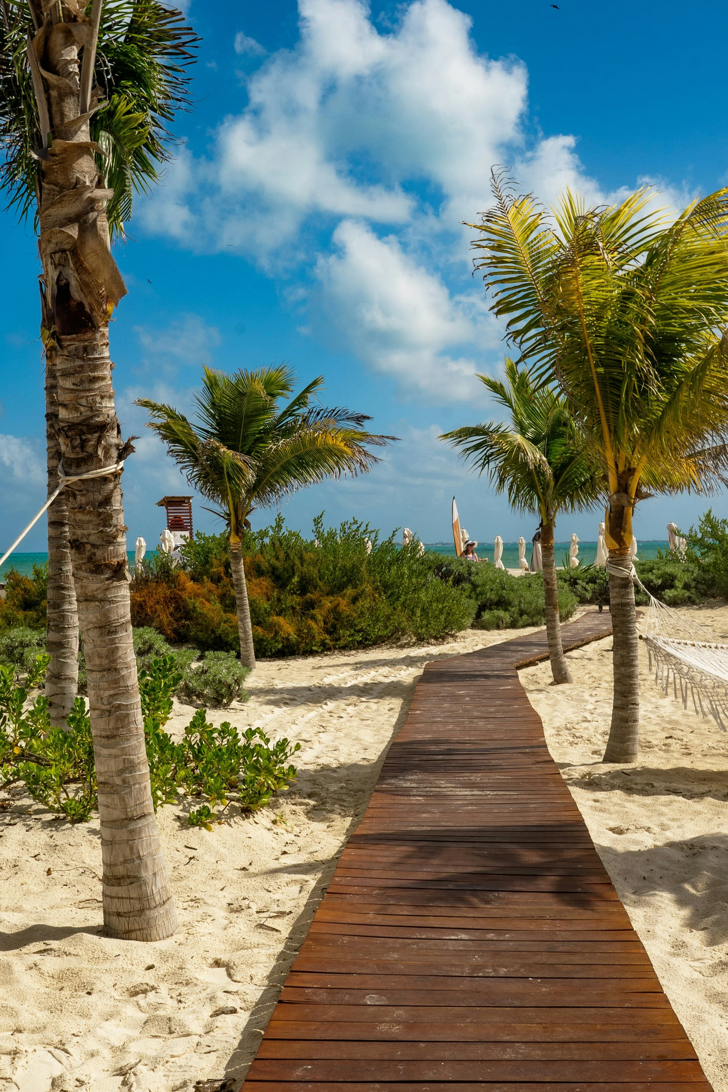 Wooden boardwalk leading to a beach with palm trees on both sides, umbrellas, and a bright blue sky with clouds.