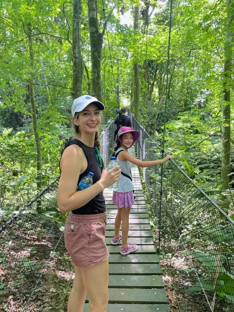 A woman and a girl holding water bottles on a suspension bridge in a lush green forest, smiling at the camera.