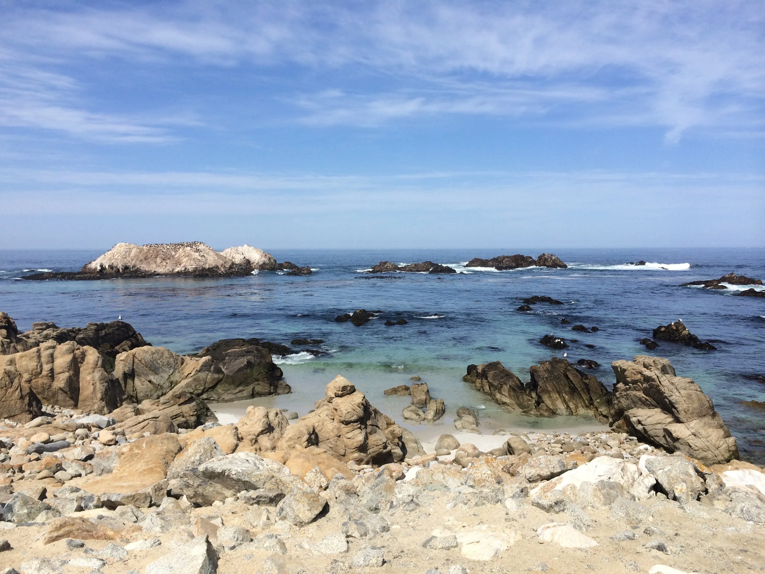 A rocky beach with large stones and pebbles in the foreground, calm ocean water with rocks extending into the horizon, and a partly cloudy blue sky overhead.