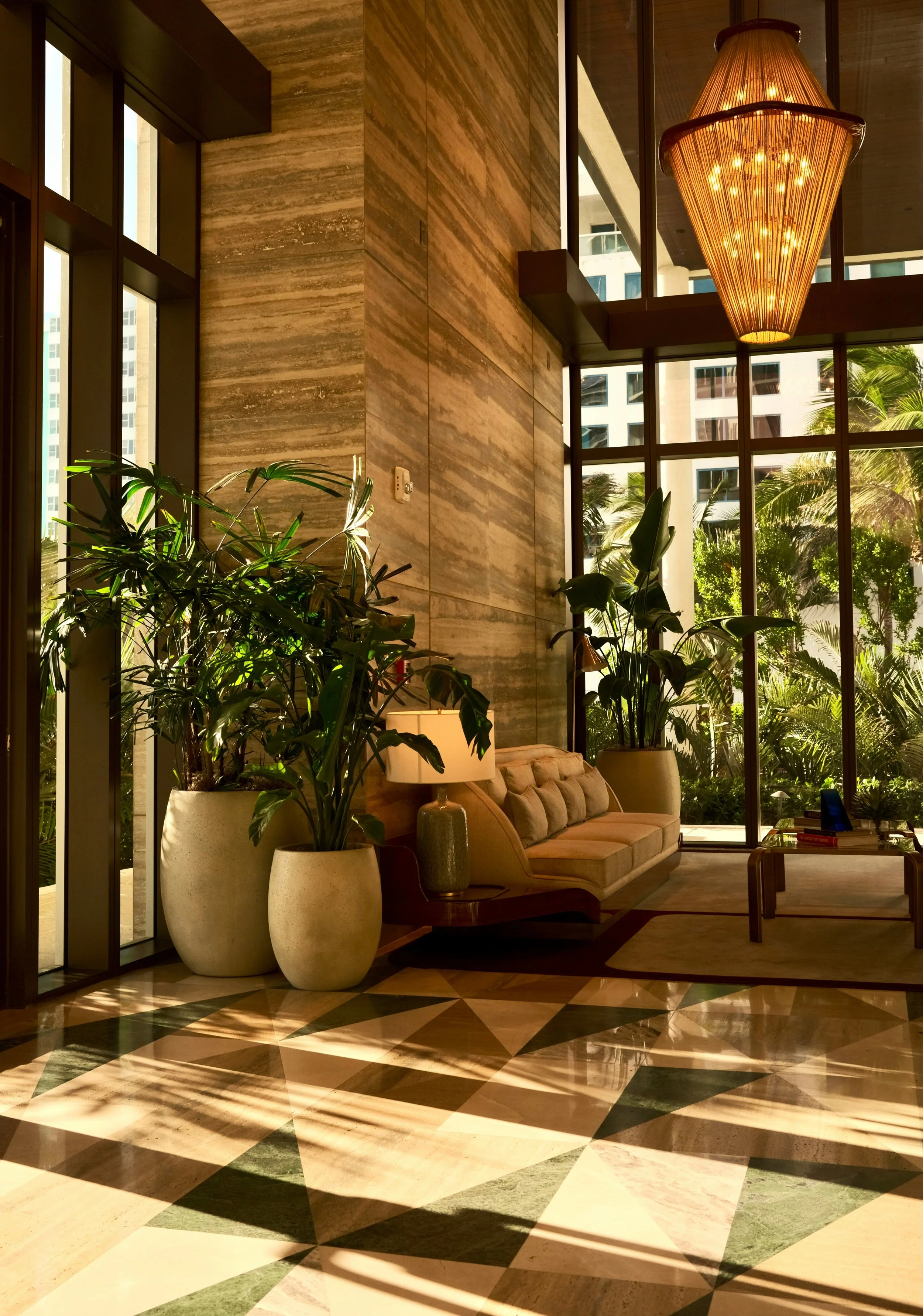 Interior view of a hotel lobby with large potted plants, a beige sofa, a lamp, and a geometric patterned marble floor. Large windows let in natural light and show a view of tall buildings and palm trees outside. A hanging chandelier is also visible.