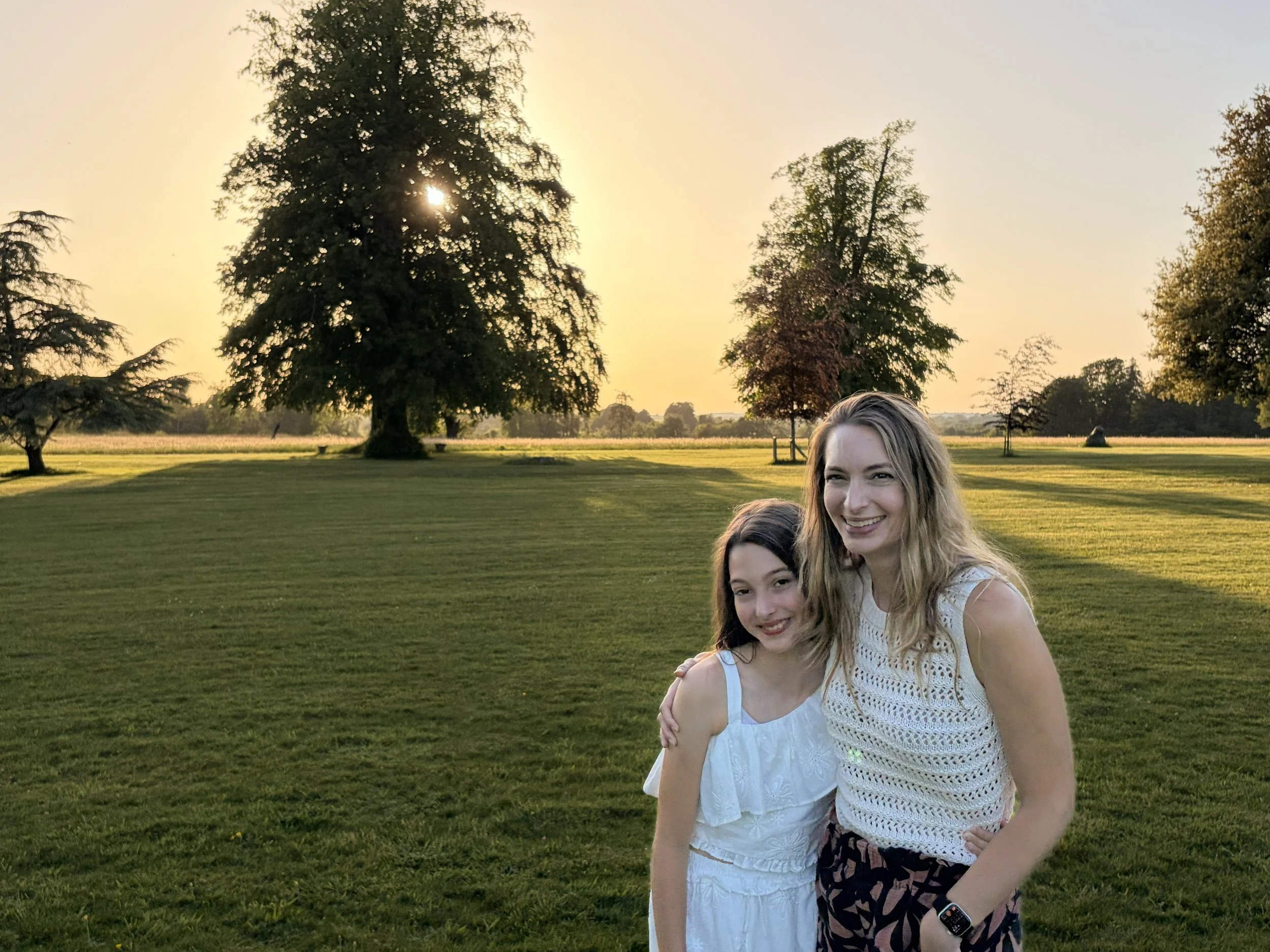 Two young women smiling in a park with tall trees and a grassy field at sunset.
