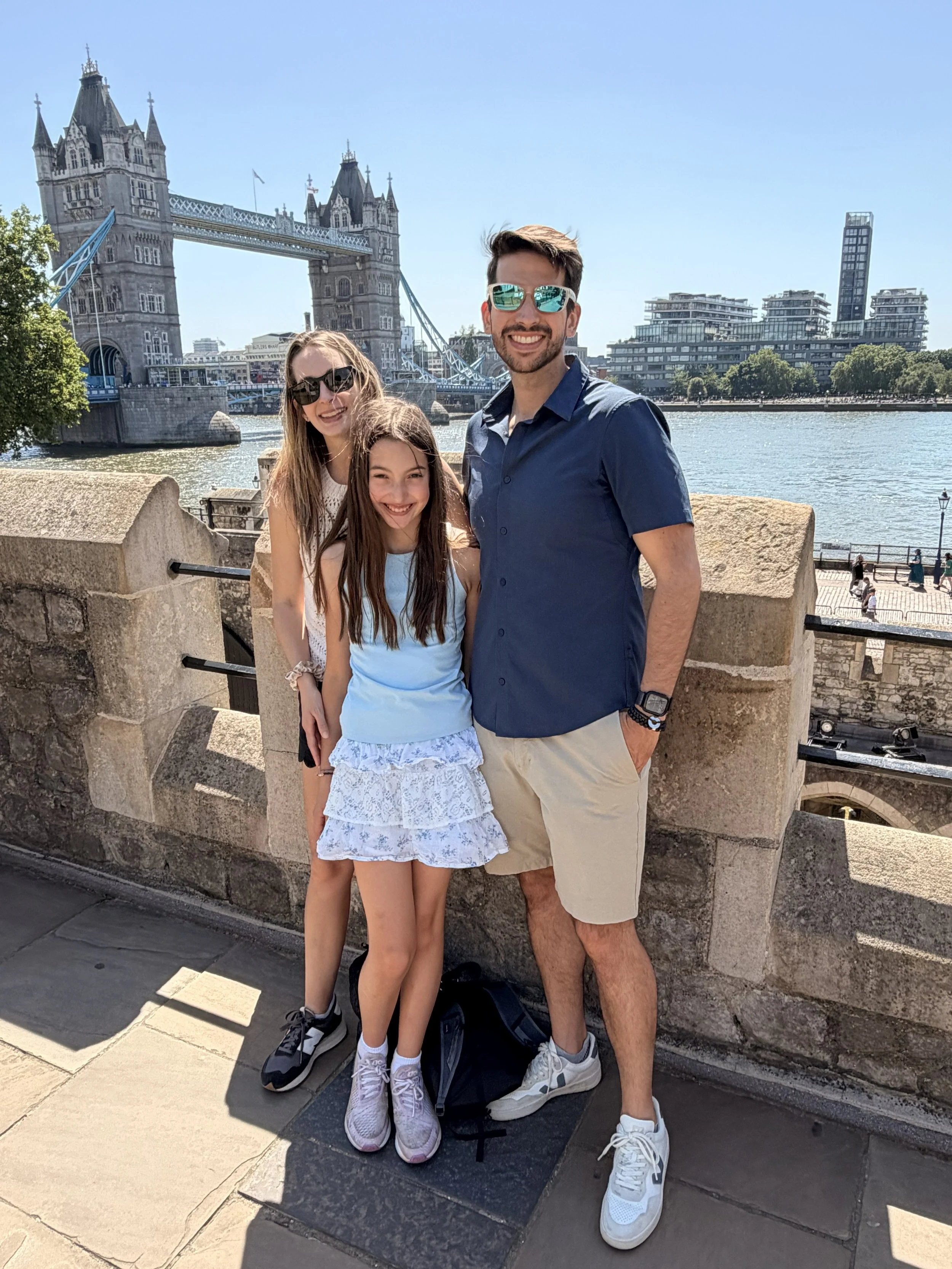 A group of three smiling people, two women and one man, standing on a bridge near the River Thames with Tower Bridge in London in the background. The man and woman are wearing sunglasses, and the women have long hair. The man is wearing a navy blue shirt and beige shorts, while the women are wearing casual summer clothes. The weather appears to be sunny and clear.