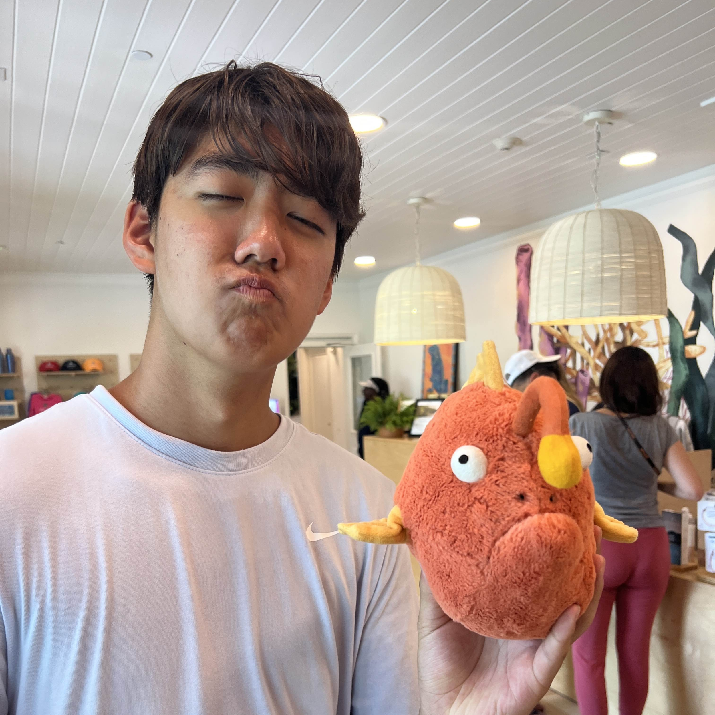 A young man with brown hair making a duck face while holding a plush toy in a brightly lit cafe.