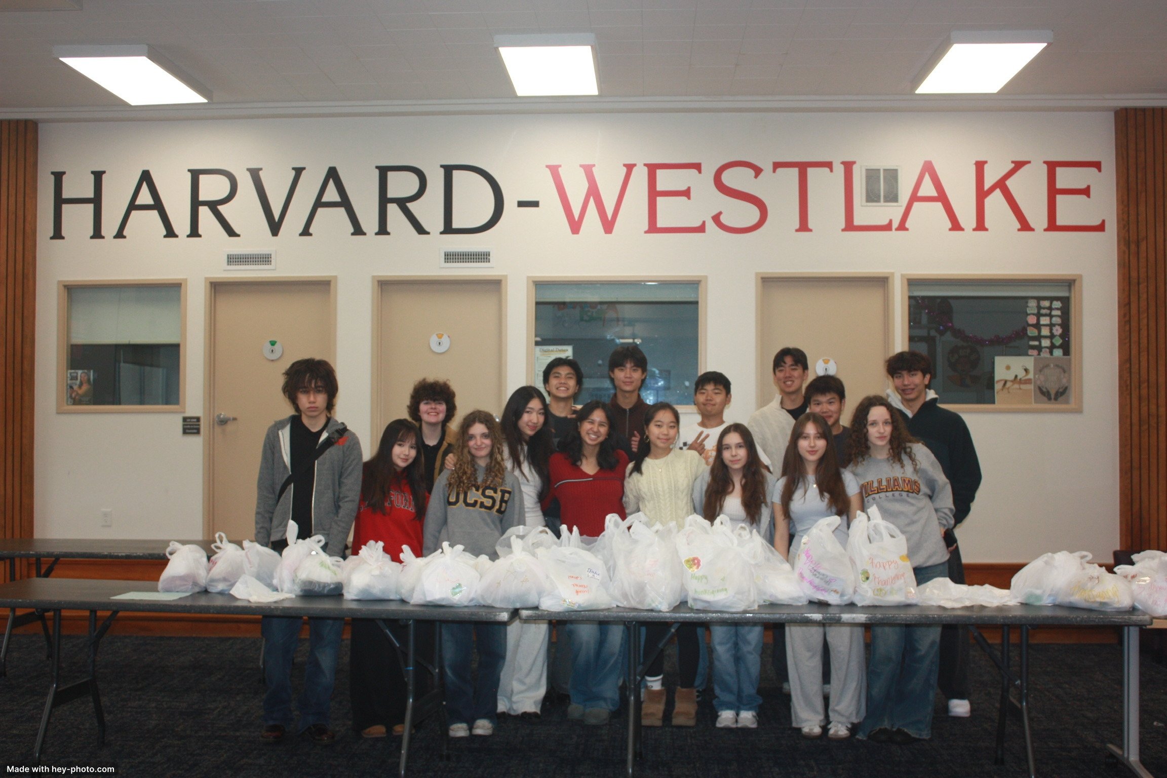 Group of students standing behind tables with bags in front and Harvard-Westlake sign on the wall.