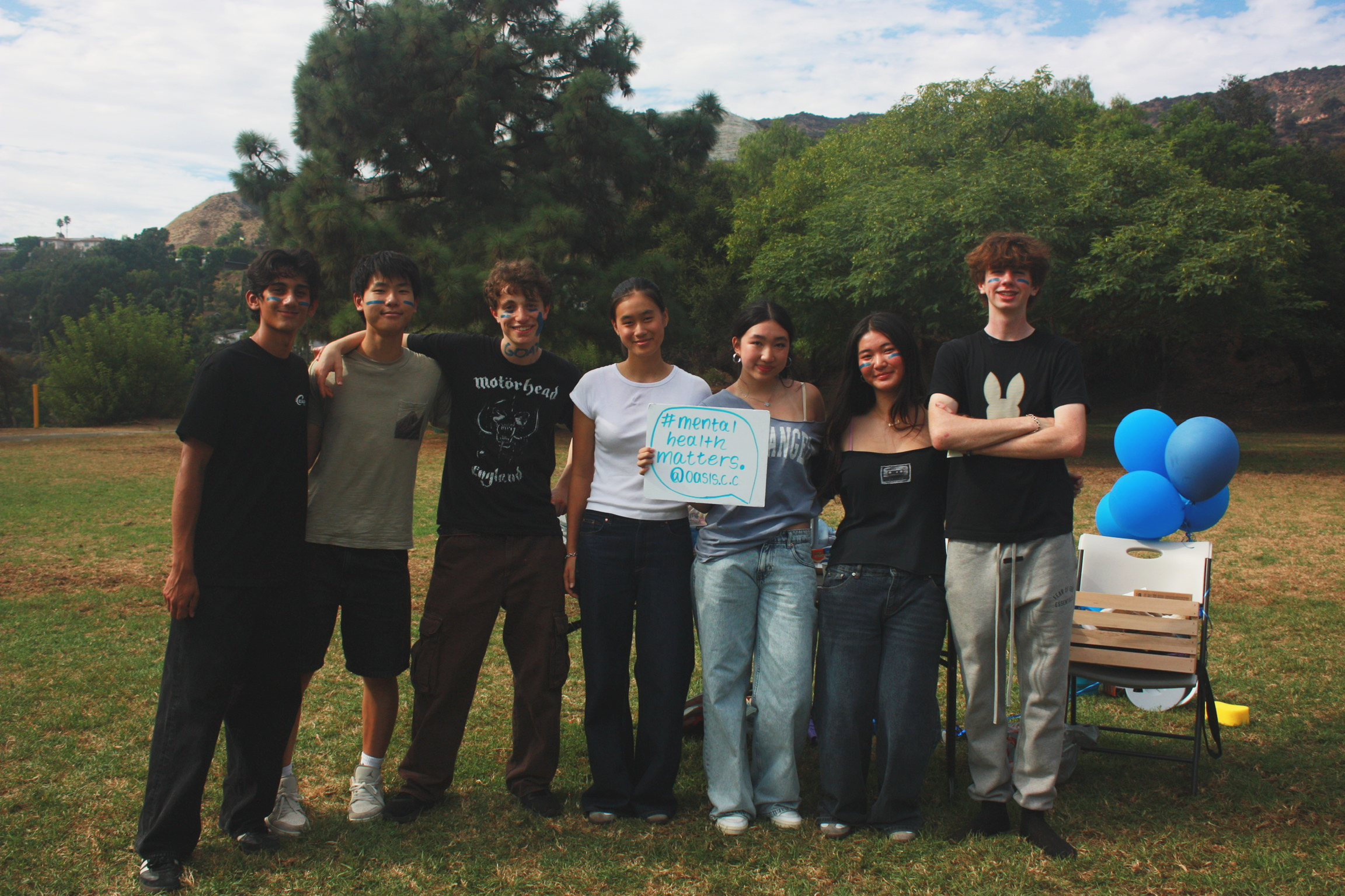 A group of eight young people, smiling and standing outdoors on a grassy field with trees and mountains in the background, participate in a mental health awareness event. One person holds a sign that says '#mental health matters. @oasis.c' and there 