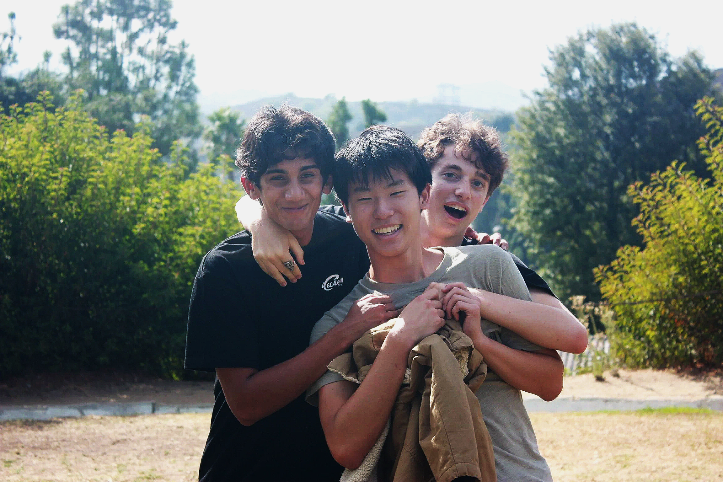 Three young people smiling and hugging each other outdoors, with trees and a cloudy sky in the background.