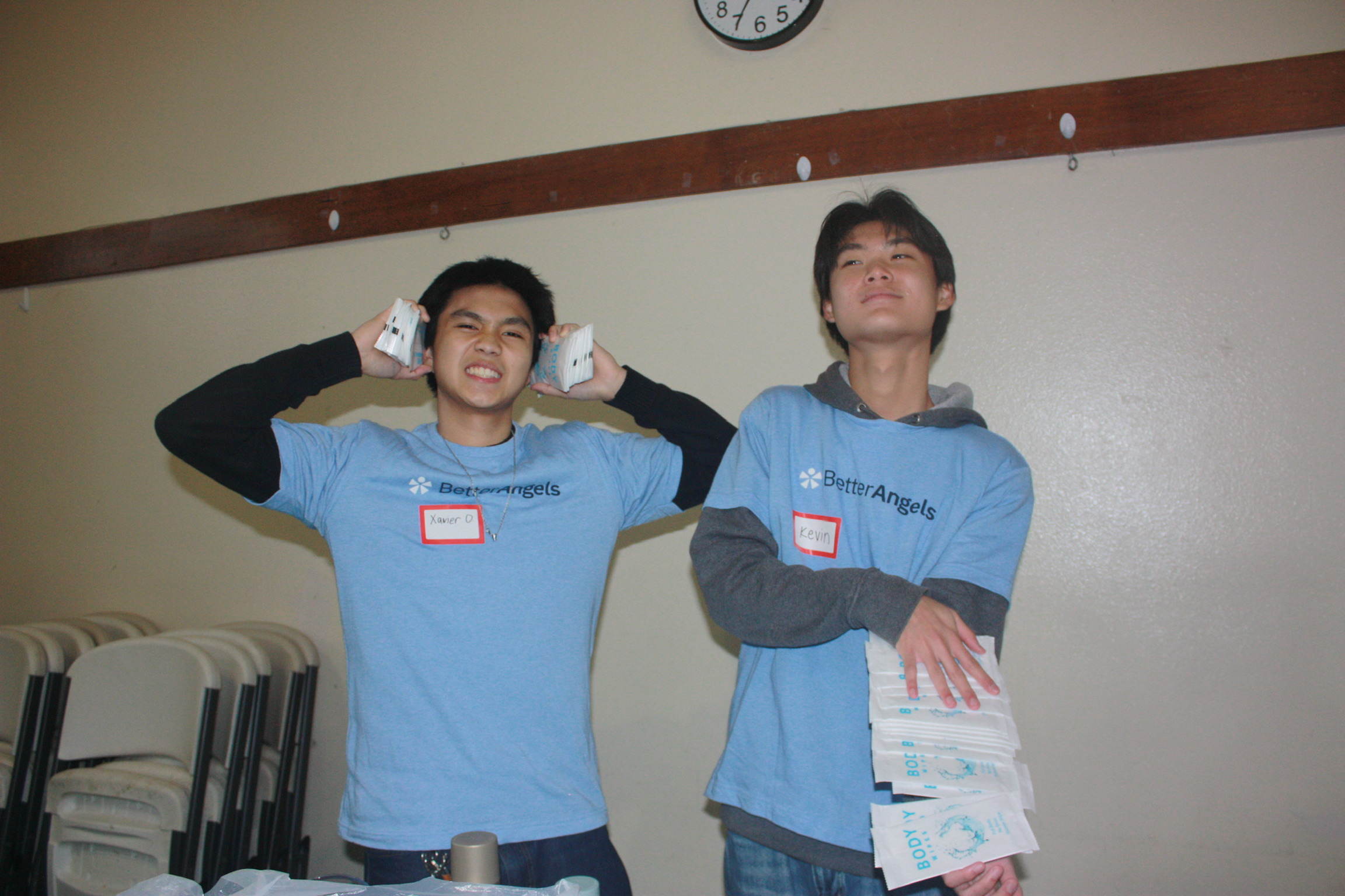 Two young men with name tags, wearing blue T-shirts, standing indoors against a beige wall with a clock, stacks of chairs, and a table with bags, one holding folded paper items.
