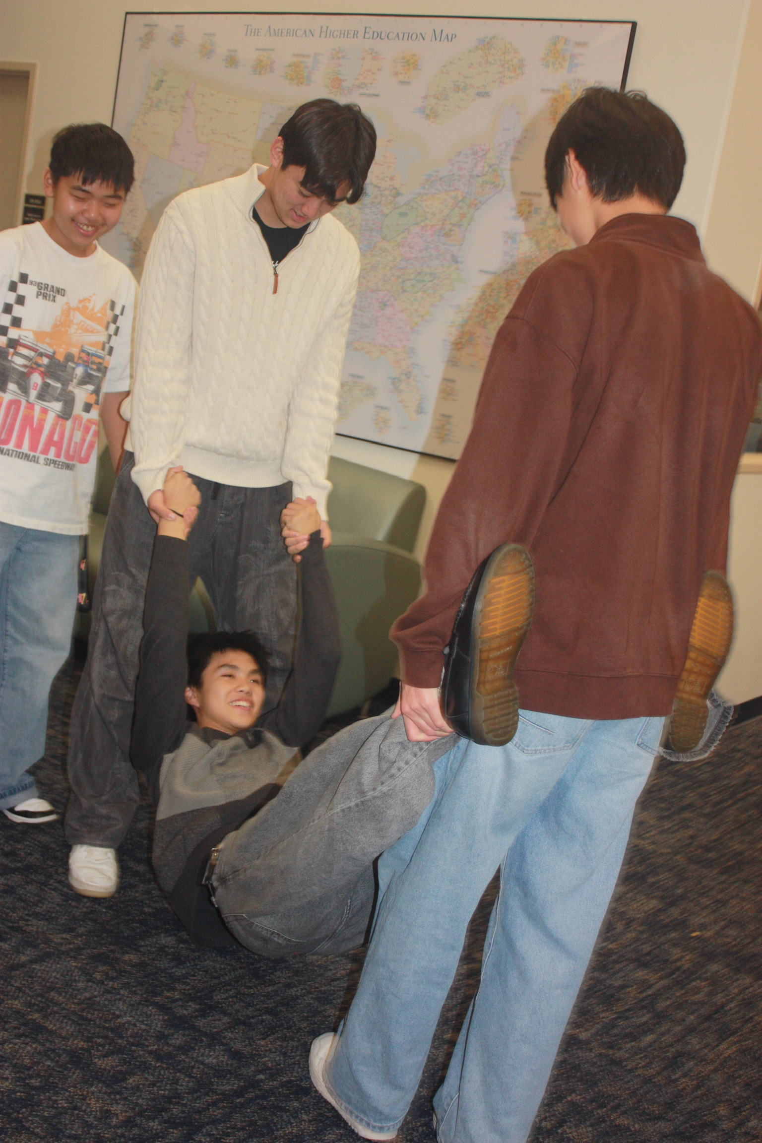 A group of young people participating in a human pretzel formation, with some standing and others on the floor, in front of a map labeled 'The American Higher Education Map.' They are indoors, in what appears to be a classroom or meeting room.