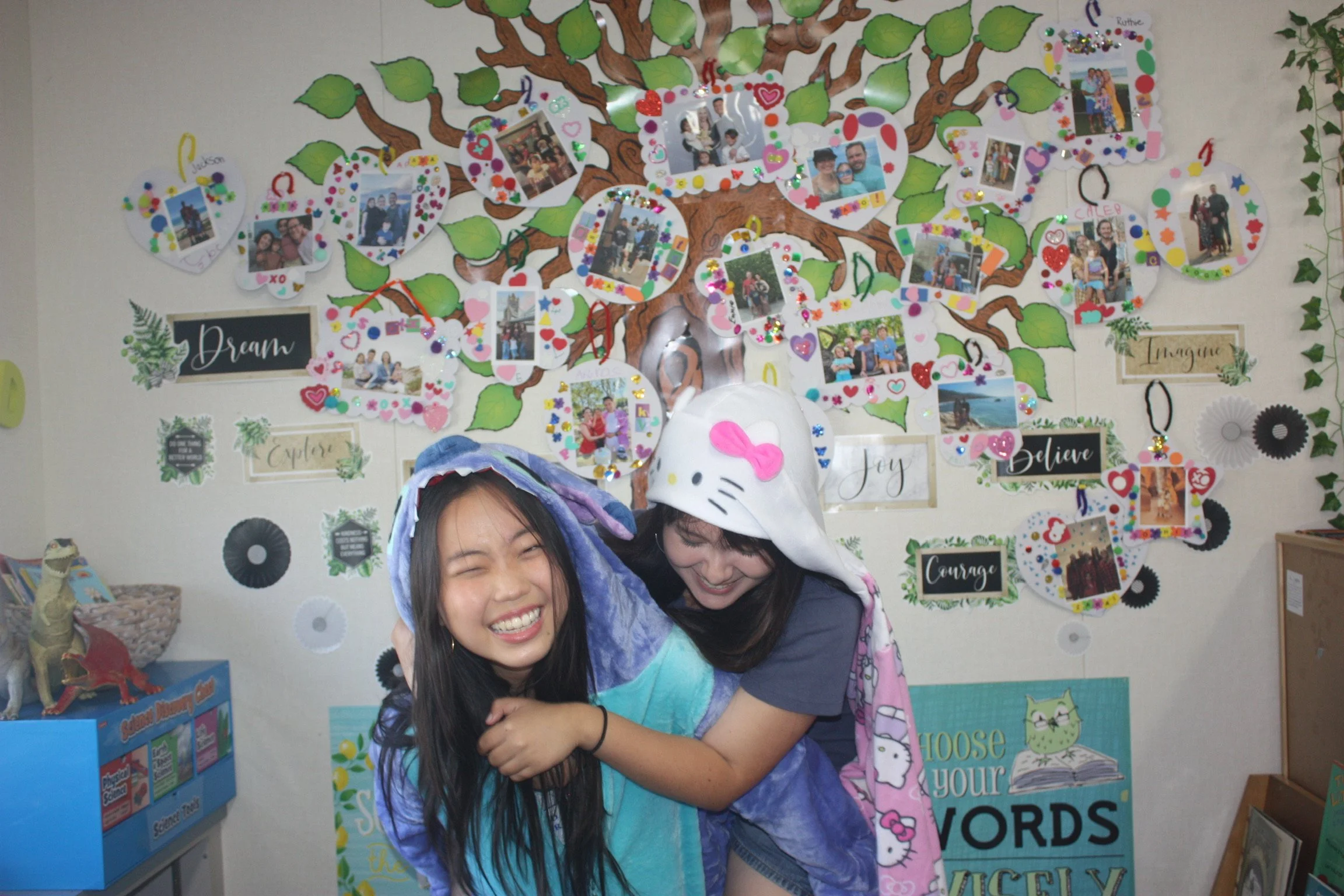 Two girls hugging and smiling in front of a colorful wall display with photos, decorations, and motivational words.
